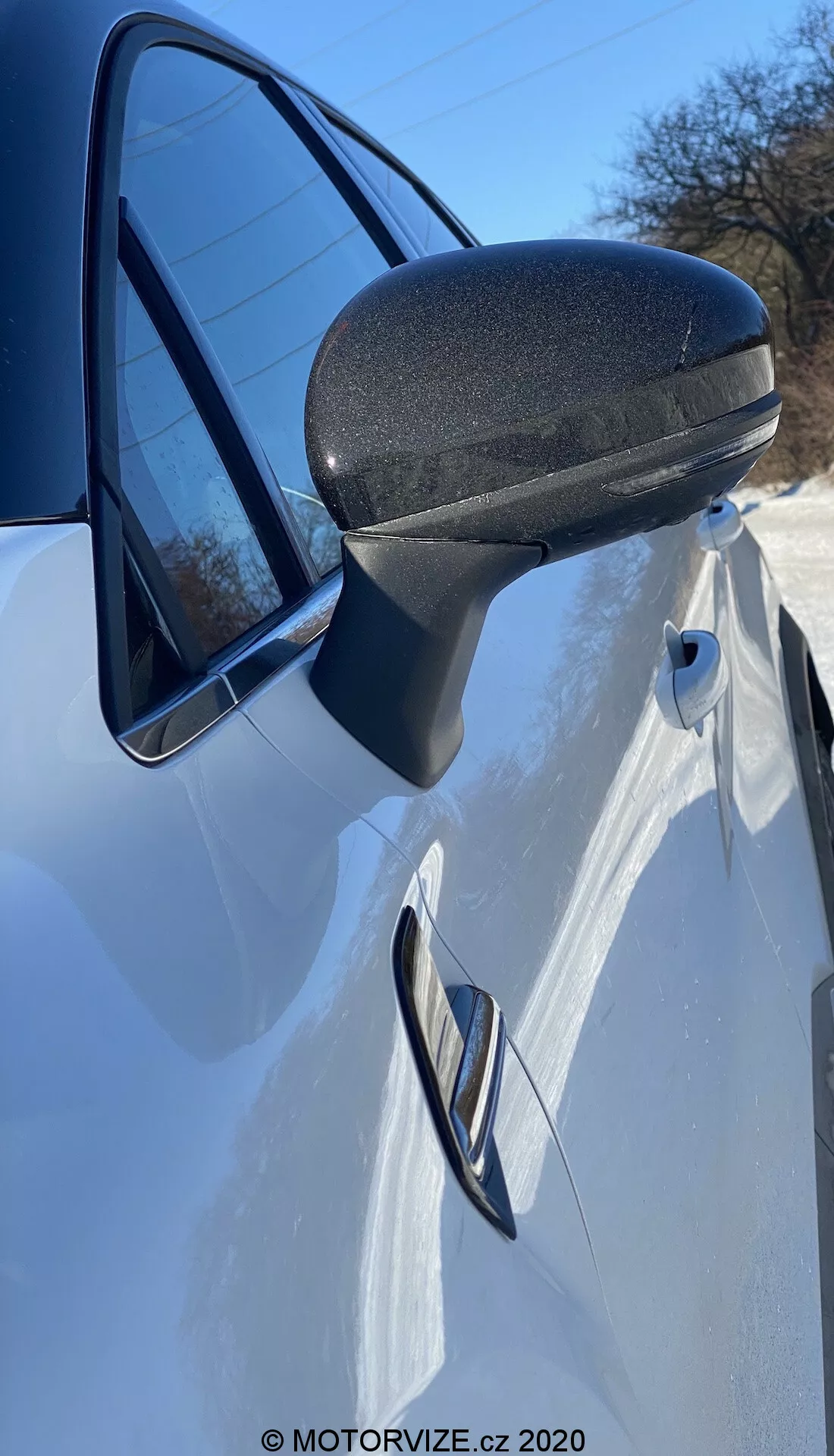 Detaljerad närbild på sidospegeln, dörren och främre kvartsdelen av en vit Renault Captur 2020. Fotot, taget från sidan precis framför sidospegeln i en uppåtvinklad vinkel, framhäver den moderna, aerodynamiskt effektiva designen av sidospegeln med en svart texturerad nedre sektion för hållbarhet. Strömlinjeformad stöd går smidigt in i dörrkarmen. Den infällda dörrhandtaget och den karakteristiska linje som går genom detta antyder en sportig, elegant design. En del av strålkastaren och en reflektion av den klara himlen i fönstret förstärker denna dynamiska presentation.
