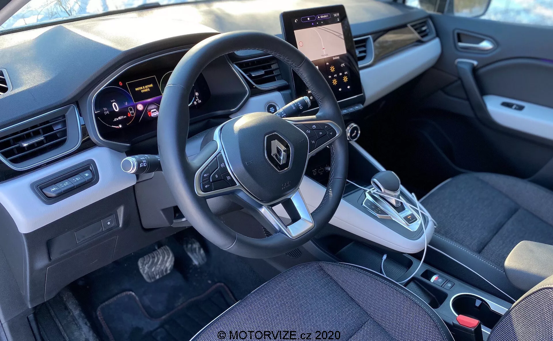 Interiörbild av en Renault Captur 2020 från baksätet, med fokus på det främre kabinområdet inklusive instrumentbräda, ratt med monterade kontroller, instrumentpanel, mittkonsol med pekskärm och klimatanläggningar, automatisk växelspak och en modern blandning av tyg- och plastmaterial.