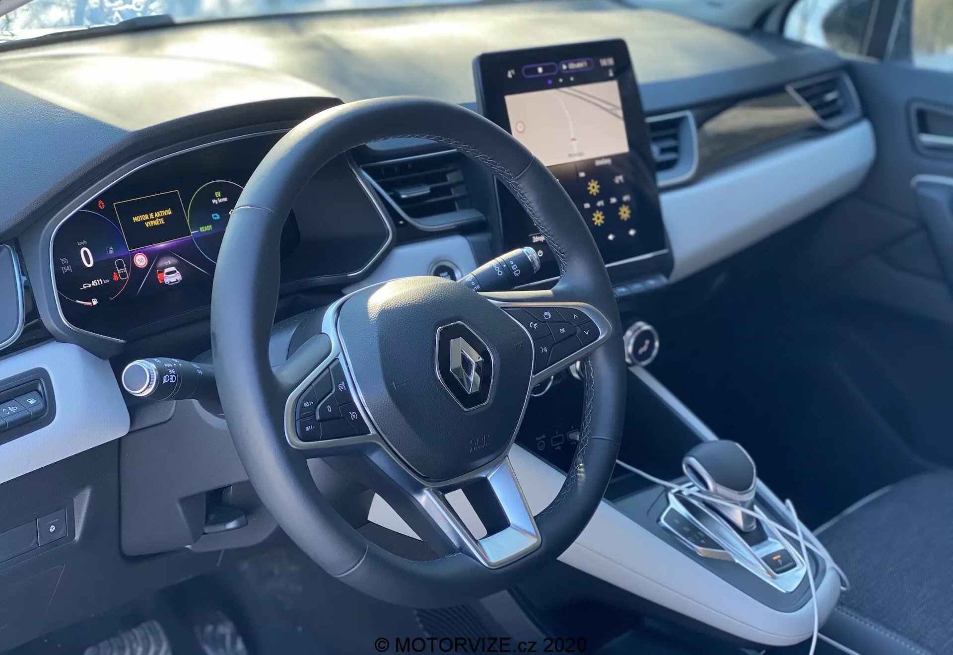 Interiör av Renault Captur 2020 från framsätet på passagerarsidan som visar den sportiga treekrade ratten med Renault-emblem och multifunktionsknappar, digital instrumentpanel med tydlig, högupplöst skärm som visar hastighet och navigationshjälpmedel, och en elegant mittkonsol. Mittkonsolen har en modern infotainmentskärm, elektronisk växelspak och en knapp för elektronisk parkeringsbroms. Den tvåfärgade grå interiören med konturerad design och accentlister ger en premiumkänsla.