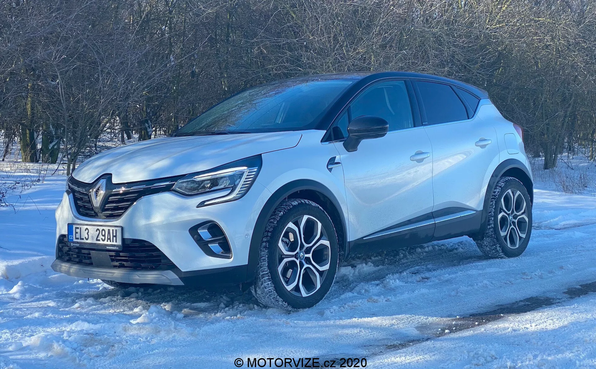 Låg vinkels vy av en Renault Captur 2020 SUV på en snötäckt yta, med frontkvartsprofil som betonas. Det starka dagsljuset och den klara himlen framhäver moderna designelement som den framträdande främre grillen med emblem, LED-dagljus, komplexa tvåfärgade lättmetallfälgar, skulpterad kaross med uttalade hjulskärmar, karossfärgade speglar med integrerade blinkers, tonade rutor och infällda dörrhandtag. Fotot betonar fordonets högkvalitativa utförande och potentiella vinterkörningskapaciteter.
