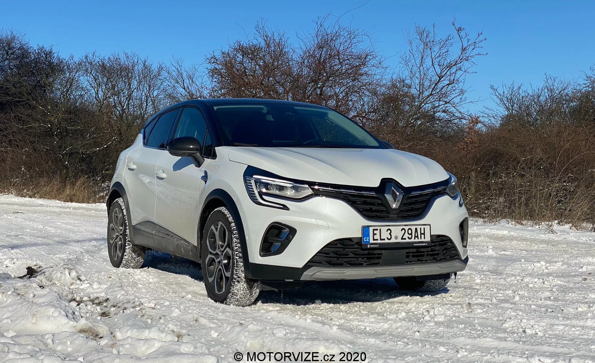Vit Renault Captur 2020 parkerad på ett snötäckt område under en klar himmel, visad från en låg, trekvartsvinkel framifrån som visar frontgrill, strålkastare med LED-dagsljus, dimljus, främre stötfångare, vinterdäck, distinkt Renault-logotyp, skulpterade linjer på huven, förhöjd markfrigång, eventuella parkeringshjälpsensorer, sidospeglar och ett övergripande rent, välskött skick, med MOTORVIZE.cz © 2020 vattenstämpel.