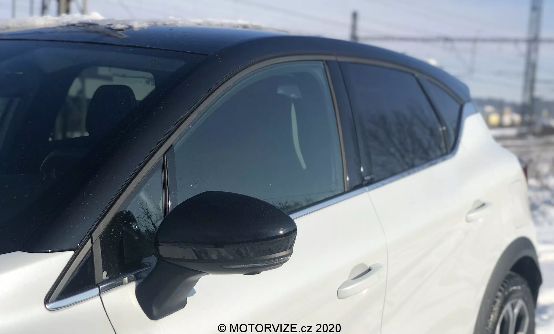 Närbild av vit Renault Captur 2020 från sidan, fokuserad från nära sidospegeln till området före framhjulet. Framträdande svart sidospegel med integrerad blinkers, fotograferad i dagsljus med naturligt solljus. Lätt lågt vinkel som betonar mittsektionen, grunt skärpedjup. Osuddad bakgrund med igenkännbara element som stängsel, kraftledningar och en struktur, vilket antyder en utomhusmiljö. Fordonets glansiga vita färg reflekterar ljuset starkt, vilket indikerar en väl underhållen modern yta. Bilens färgmatchade dörrhandtag och stigande fönsterlinje mot bakre delen visar en dynamisk design.