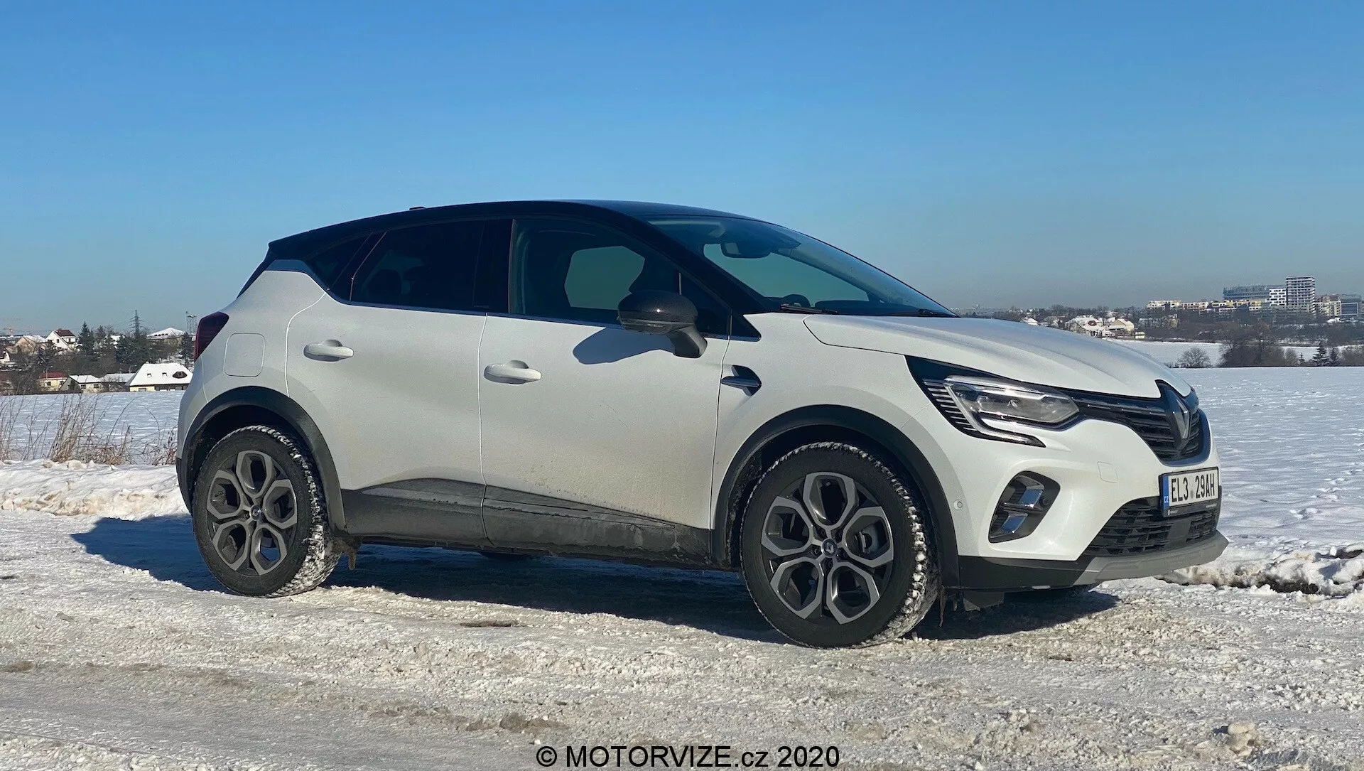 Renault Captur 2020 modern crossover SUV placerad på isigt och snöigt terräng med klar himmel i bakgrunden. Passagerarsidan vetter mot kameran i en lätt vinkel, vilket visar fronten, sidan och delvis ovansidan av fordonet. Funktioner inkluderar LED-dagljus, integrerat frontgaller med tillverkarens emblem, tvåfärgad ljus och svart lack, femekrade lättmetallhjul, plastskyddslister, eleganta infällda dörrhandtag och en avsmalnande fönsterlinje med bakspoiler. Föreslår avancerad teknik som LED-belysning och nyckelfritt intåg, satt i ett kallt väderlandskap nära ett stadsområde.