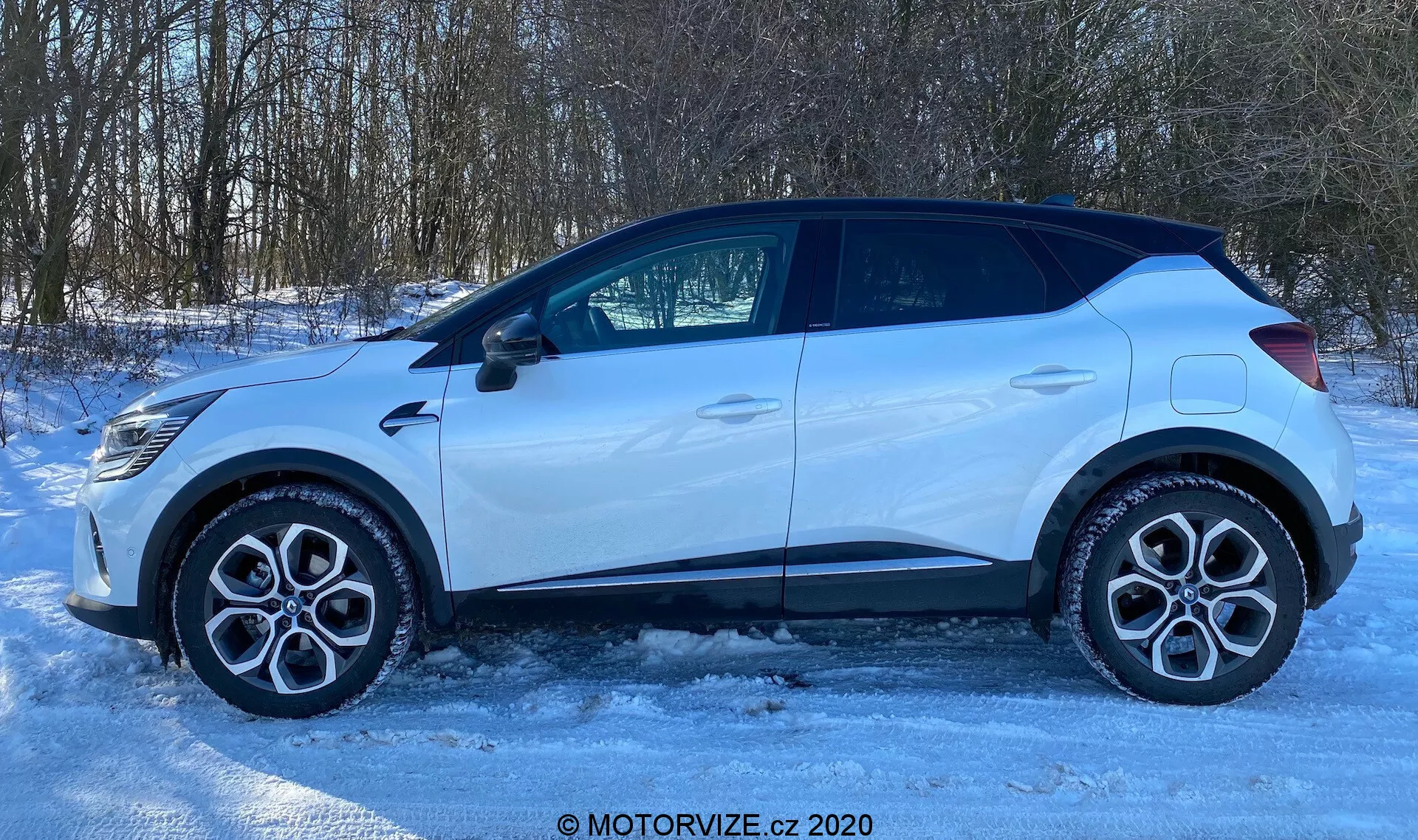 Sidovy av en Renault Captur 2020 parkerad på en isig eller snötäckt yta. Bilens profil visar lättmetallfälgar, standardvägdäck, fyra dörrar, sidofönster inklusive ett litet bakre fyrkantsfönster och en sidobackspegel med en integrerad blinkersindikator. Taklinjen lutar något mot baksidan, med plastklädsel runt hjulhusen och den nedre delen av dörrarna, vilket antyder en terrängklar design. Den välbelysta scenen, troligen från naturligt solljus, definierar bilens form mot en lantlig eller förortsmiljö med träd i bakgrunden. Bilden har en vattenstämpel med texten 'MOTORVIZE.cz 2020'.