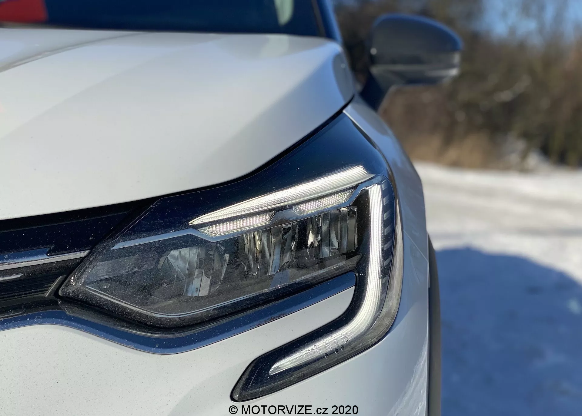 Närbild av vänster främre hörn på en modern bil, med fokus på LED-strålkastare och grill, tagen från en låg vinkel nära strålkastarnivån. De dagljuslamporna (DRL) har en kurvig design som omfamnar strålkastarenheten. Potentiellt adaptiva strålkastare eller hörnljus synliga, med metallisk lack och integrerade blinkers i ytterbackspegeln. Ljus naturlig belysning antyder en utomhusdagmiljö.