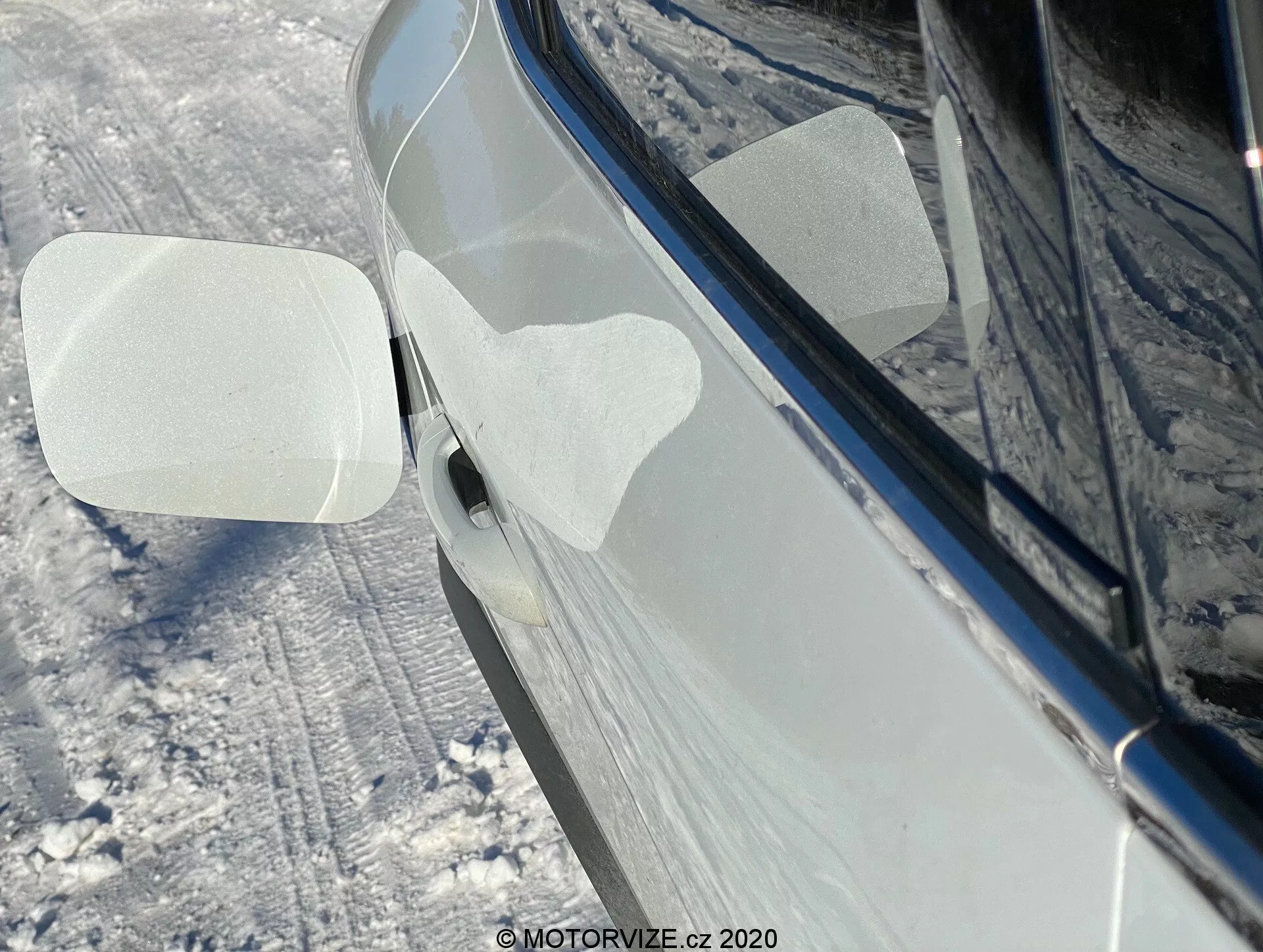 Närbild av sidan på en 2020 Renault Captur i en snöig utomhusmiljö. Fokuset ligger på höger sidospegel, som är vikt framåt, vilket kan indikera en automatisk eller manuell fällningsfunktion. Spegelns hölje har två transparanta skyddsfilmer fästa i form av parallellogrammer, sannolikt för skydd under leverans eller tvätt. Bilen, som verkar ha en mörk, blank nyans som svart eller mörkblå, ser väl underhållen ut med reflektioner som tyder på en ren, polerad yta. Marken runtomkring och sidorna av bilen är täckta av snö. En del av det främre dörrhandtaget med en knapp för nyckelfri inpassering eller beröringssensor är också synlig.