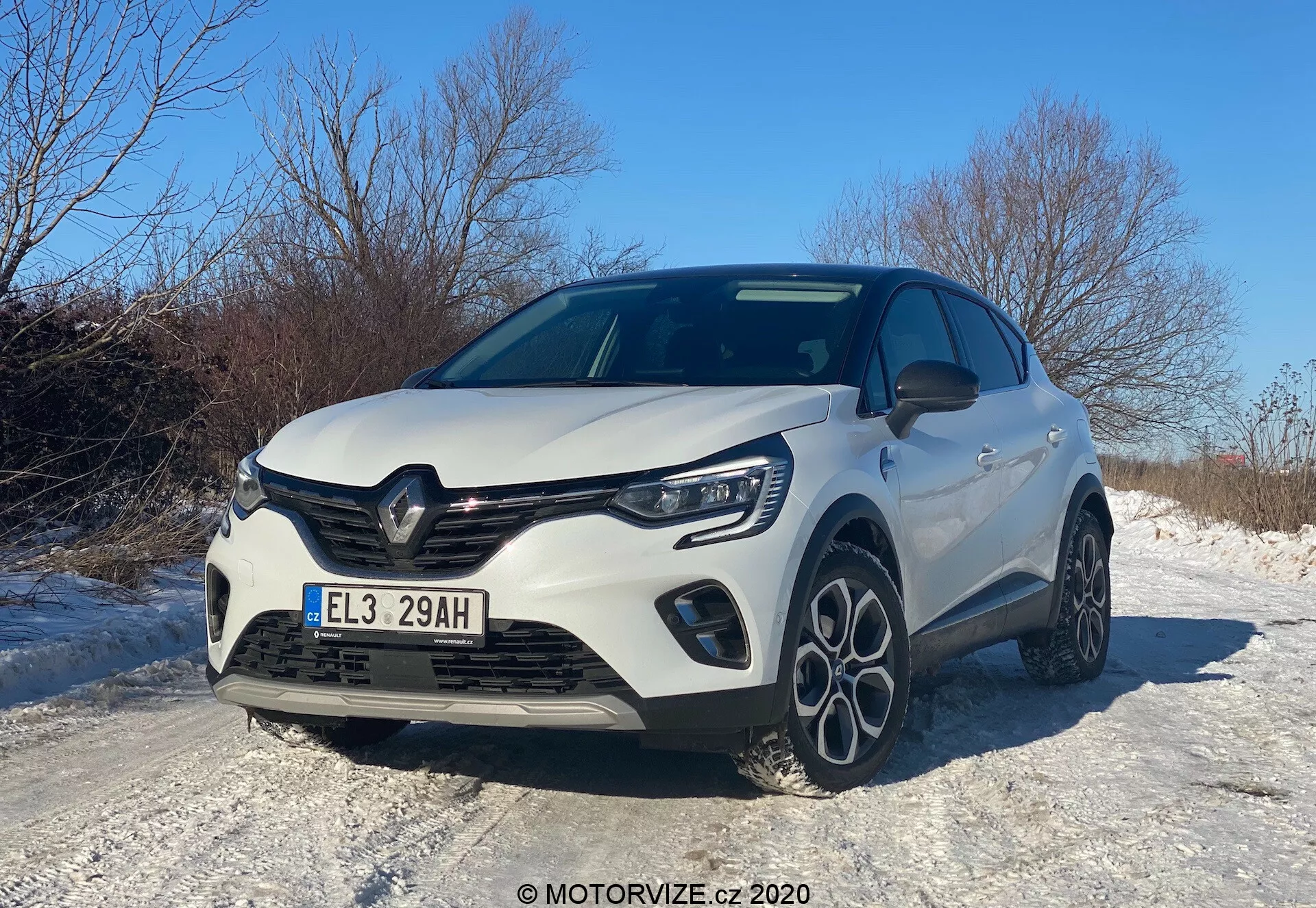 Modern hatchbackbil Renault Captur 2020 fångad från en låg framifrån trekvartsvinkel, som visar bilens främre grill, motorhuv, LED-strålkastare, främre stötfångare med integrerade dimljus och delvis sidoprofil. Designelementen inkluderar en framstående grill med tillverkarens emblem, skulpterade linjer på motorhuven och flerspakslegeringshjul, allt under en klar daghimmel, vilket reflekterar en sportig och aerodynamisk estetik, möjligen framhävande en uppgraderad trim- eller sportpaket. Bilen är placerad på en snötäckt väg, vilket antyder egenskaper som är lämpade för vinterkörning.