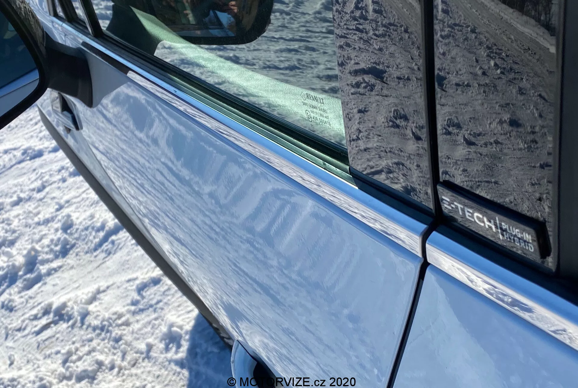 Närbild av Renault Captur 2020 i metallicblått med fokus på förardörren och sidospegeln med integrerad växelsignal. Bilden visar dörrområdet med en svart kant längs kanten av det tonade fönstret, som reflekterar den omgivande miljön. På dörren syns delvis en logotyp som indikerar en möjlig utrustnings- eller teknologipaket.