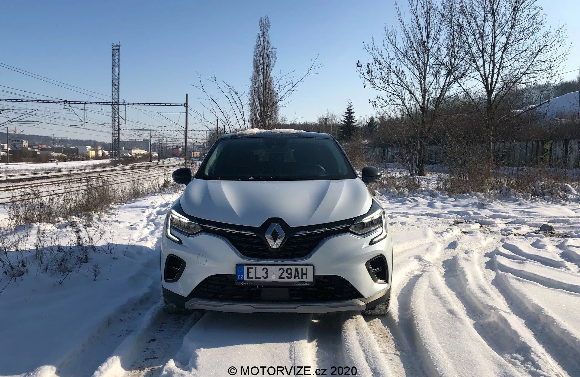 Rakt på framifrån vy av Renault Captur 2020 på en snötäckt väg, som detaljerat visar främre grillen, strålkastarna, skulpterad motorhuv och främre stötfångare under dagtid. Däckspår är synliga och visar på nylig rörelse. Fordonets design antyder aerodynamisk stil, med framträdande varumärkesemblem och avancerat belysningssystem. Möjliga förarassistanssensorer eller kameror kan vara integrerade i främre stötfångaren.