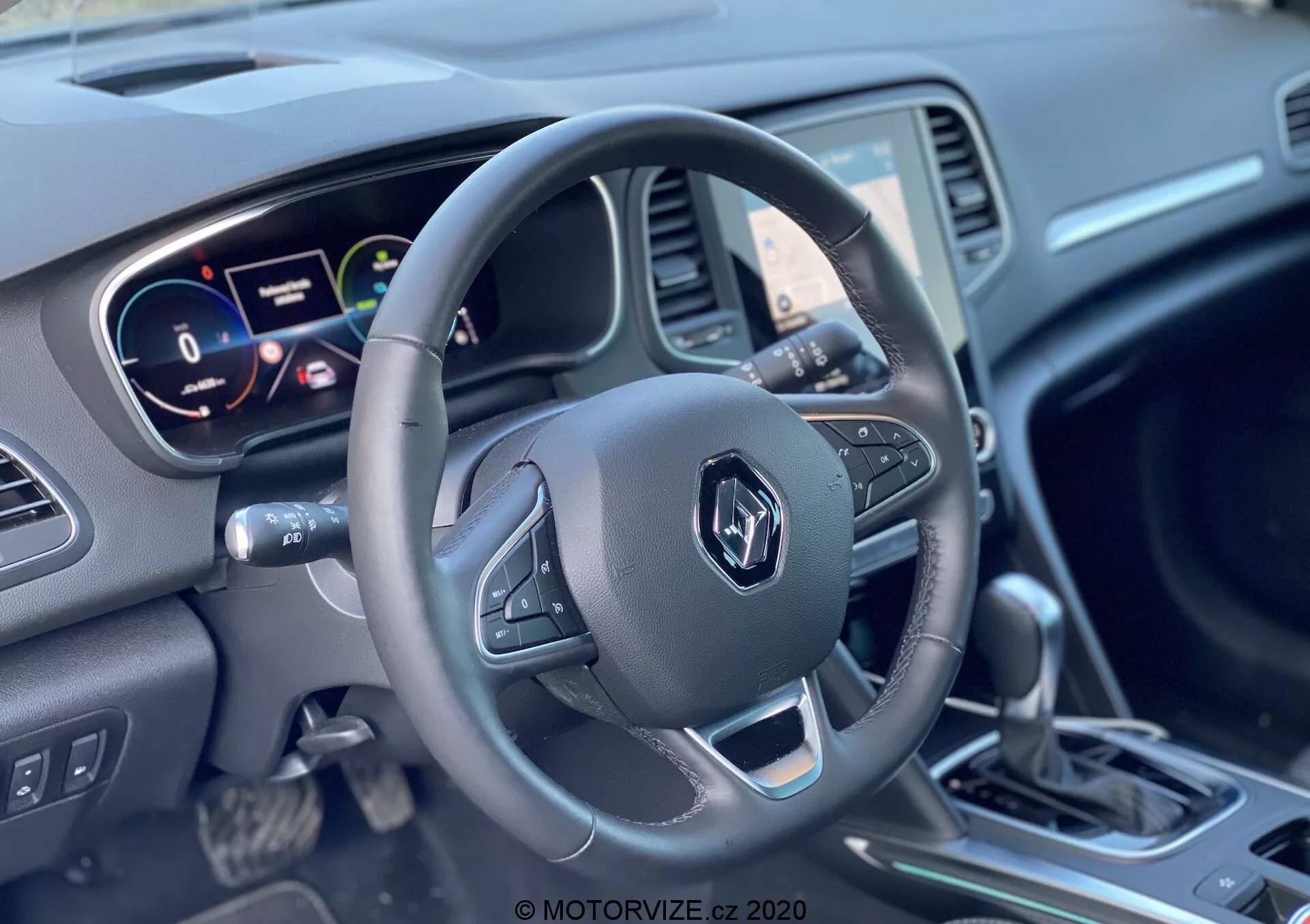 Interiörvy av en Renault Megane E-Tech 2022 med fokus på förarens perspektiv på rat och instrumentpanel, som visar en treekrad ratt med kontrollknappar, en digital och analog instrumentkluster samt en växelspak omgiven av olika kontroller.