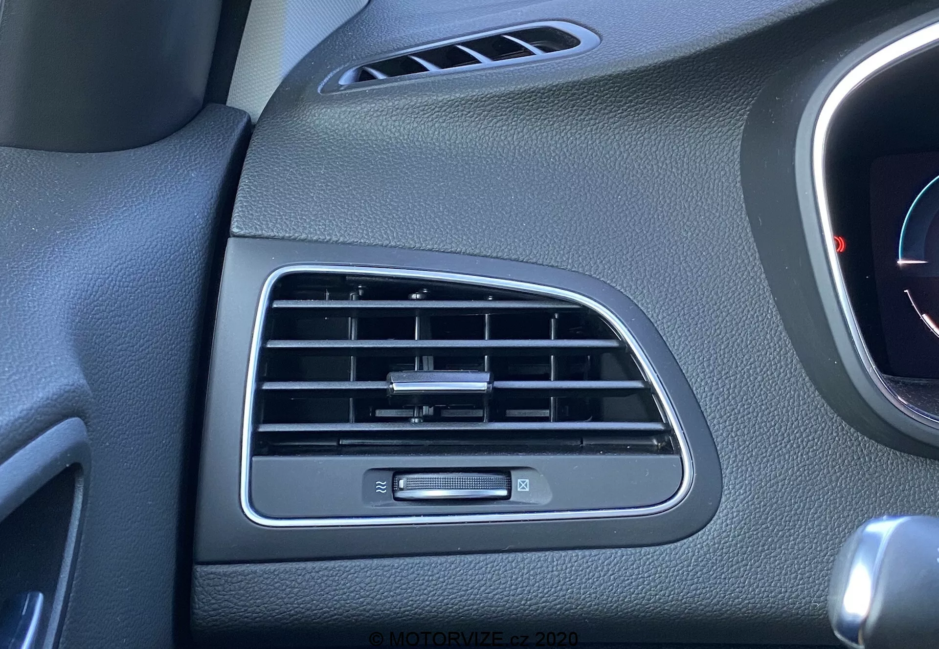 Närbild av vänstra luftkonditioneringsventilen och kontrollknappen på Renault Megane 2022 E-Tech:s passagerarsida av instrumentbrädan. De svarta lamellerna på ventilen är horisontellt orienterade, med en liten knapp på den vertikala axeln för att justera luftflödet. Till höger syns en liten del av ett oidentifierat mätinstrument eller panel. Instrumentbrädans ytor har en svart plaststruktur som är upplyst av dagsljus.