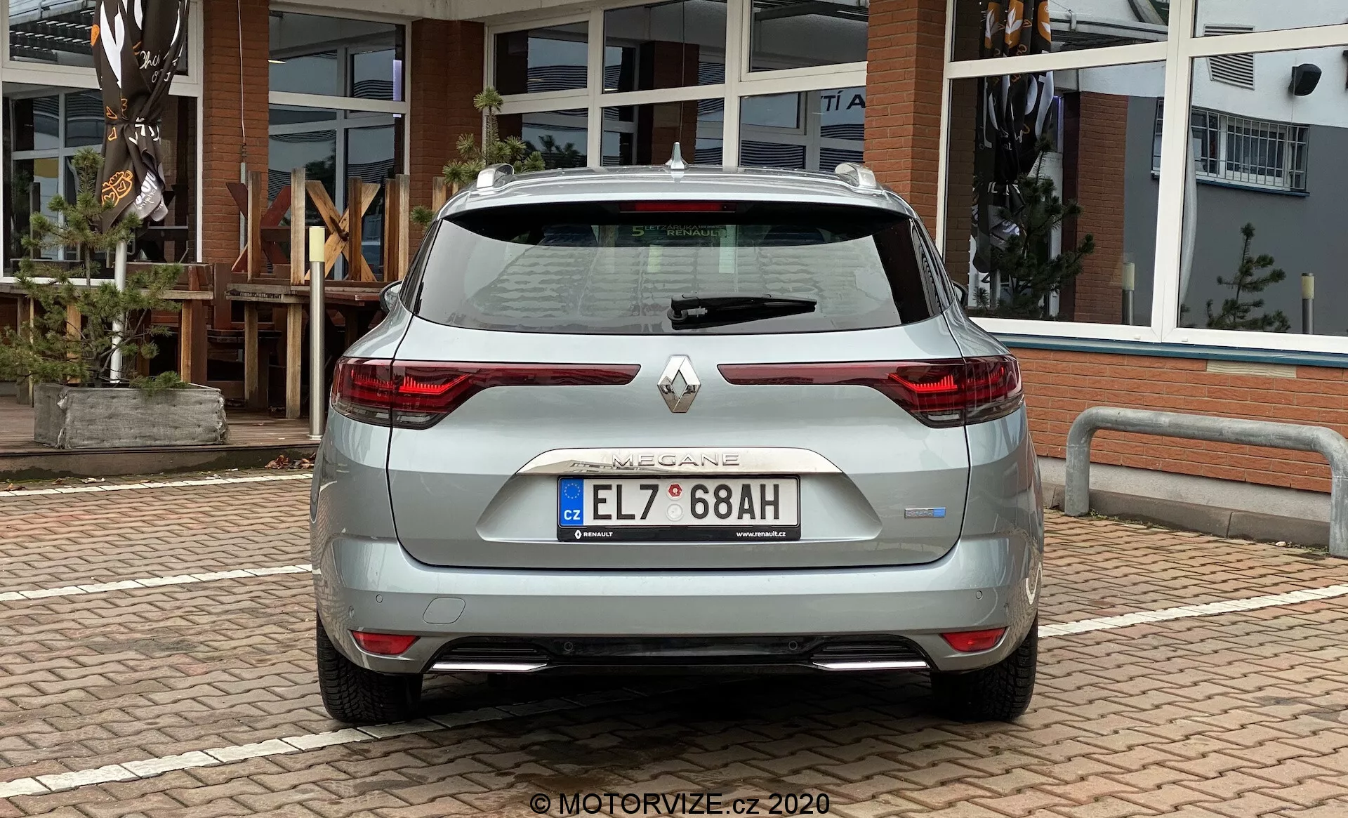 Bakre vy av en ljusfärgad hatchback-bil tillverkad av Renault, som har en baklucka över hela bredden med integrerad spoiler, moderna C-formade LED-bakljus, parkeringssensorer, backkamera, dubbelt avgasrör och kromdetaljer på bakre stötfångaren. Fordonet är parkerat utomhus på ett asfalterat område med en byggnad med glasdörrar och fönster i bakgrunden.