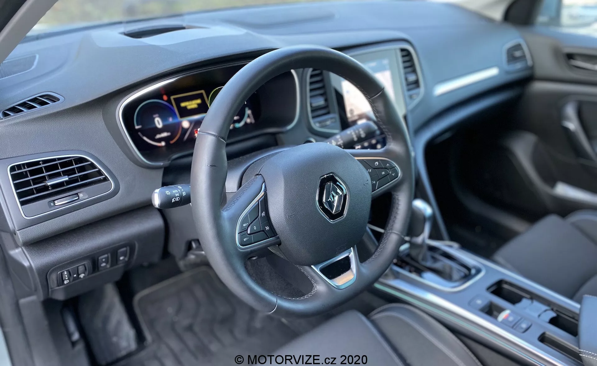 Bilden visar interiören på en Renault Megane E-Tech 2022 från förarsätet. Fotat från en något avvikande vinkel, betonar den ratten med multifunktionsknappar, en delvist synlig digital instrumentpanel bakom som visar färgglada mätare, samt ett infotainmentsystem med skärm och knappar under i mittkonsolen. Mittkonsolen har en växelspak, vilket indikerar en automatisk växellåda och olika kontroller. Interiören har en blandning av material inklusive metalliska detaljer och mjuka ytor. Väl upplyst, bilden är skarp på ratten och mittkonsolen, med ett vattenmärke från MOTORVIZE.cz 2020 i botten.