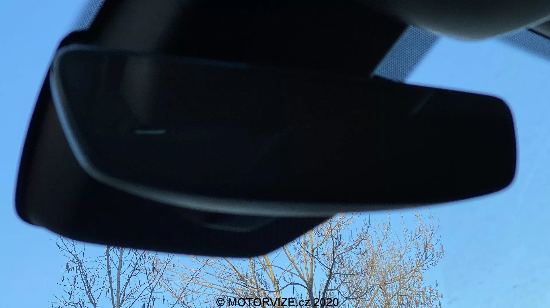 En unik interiörvy av Renault Megane 2022 (Megane-e-Tech) med fokus på backspegeln från en låg vinkel. Spegeln är centrerad, inramad av en klarblå himmel och nakna trädgrenar, vilket framhäver dess moderna, strömlinjeformade design.