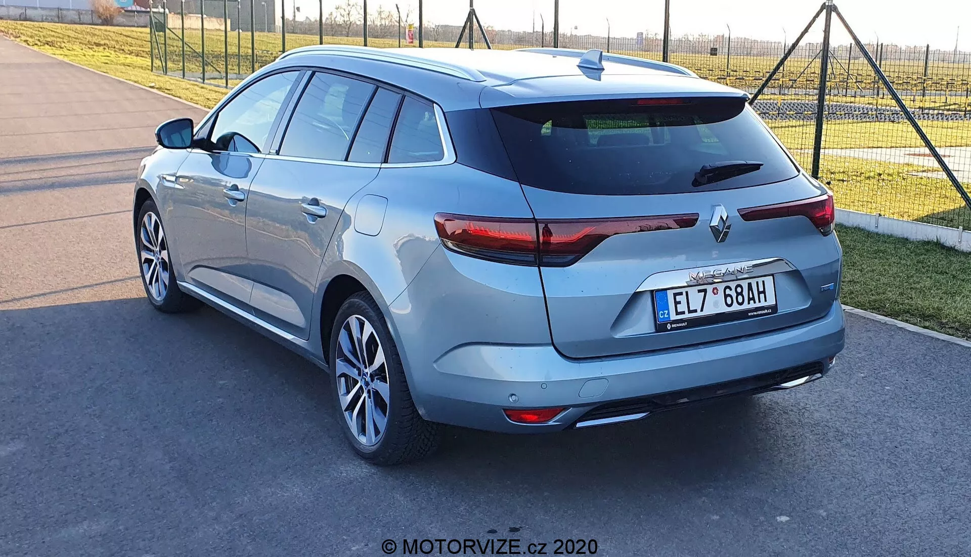 Trebildsvy bakifrån på en Renault Megane-e-Tech hatchback 2022, fotograferad under dagtid från låg till medelhög höjd, fokuserad på fordonets högra sida och bakdel. Funktioner: lutande bakruta med takspoiler, LED-bakljus, enkel avgasrör, lättmetallfälgar, bakrutespolare, parkeringskamera bak och ultraljuds sensorer för parkering. Den metalliska lacken lägger till visuell attraktionskraft, med den europeiska mittmonterade registreringsskylten som ytterligare framhäver denna väl underhållna eller nya bil.