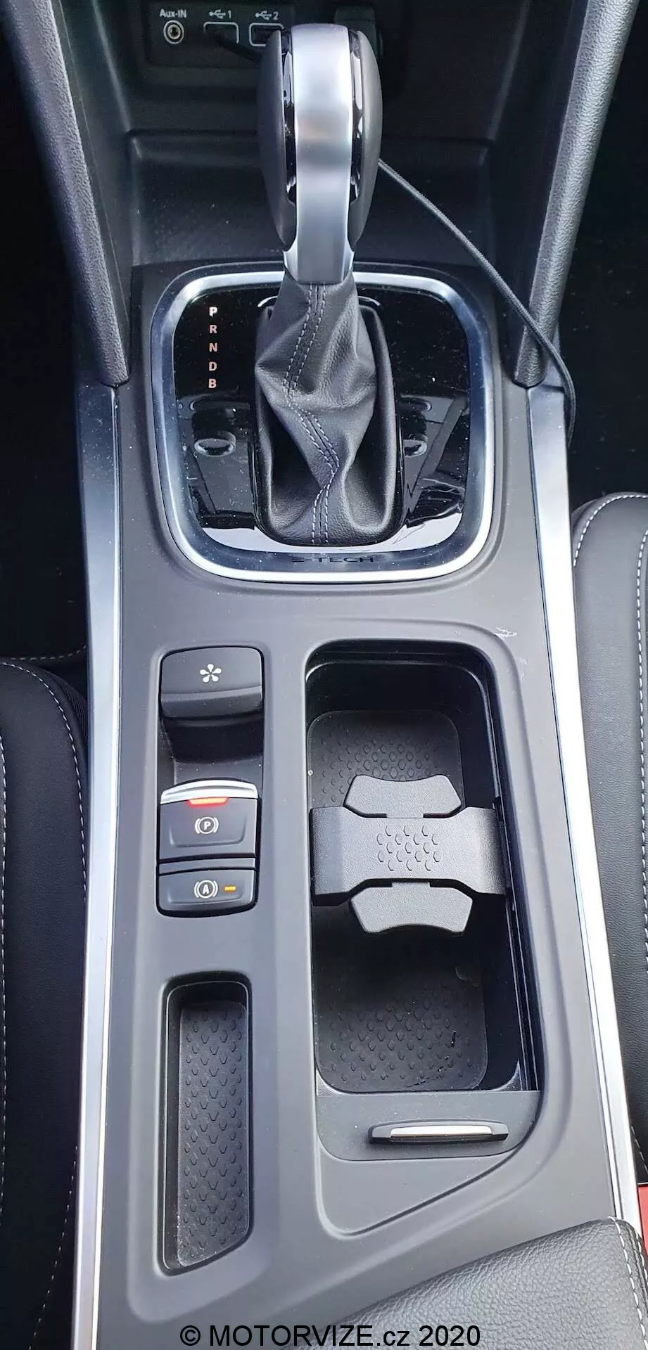 Närbild, vertikal bild av mittkonsolen i Renault Megane E-Tech 2022 sett ovanifrån med en svagt framåtlutad vinkel. Visar en automatisk växelspak omsluten av läder med silverbas i 'park' läge, högblank svart ram med upplysta PRND-indikatorer, texturerad yta bakom växelspaken, elektronisk parkeringsbroms och Auto Hold-knappar, dubbla mugghållare med en nyckel i en av dem, samt en blandning av glansiga och texturerade material med metalliska accenter.