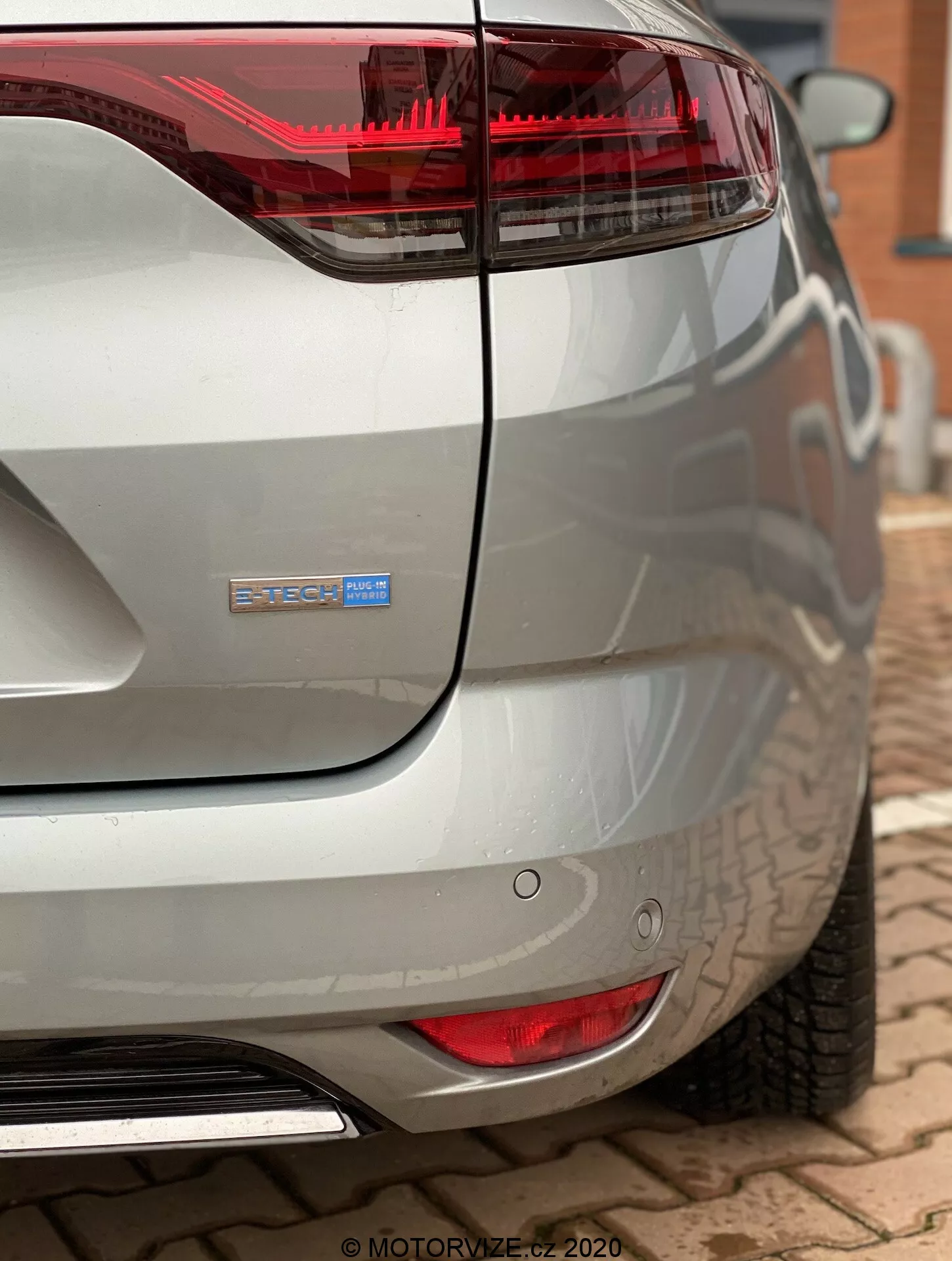 Närbild av den bakre hörnet av en modern Renault Megane 2022 (Megane-E-Tech) tagen från en låg, bakåtvänd vinkel. Bilden framhäver den röda och genomskinliga baklyktan som sträcker sig in i den eleganta bakre kvartspanelen, en försiktigt böjd bagagelucka med en suddig badge för integritet som antyder en hybrid eller elektrisk variant. Synliga funktioner inkluderar runda parkeringssensorer under baklyktan, en blank metallisk avgasrör integrerad i den böjda bakre stötfångaren och en metallisk silver, reflekterande lack som fångar omgivande föremål.