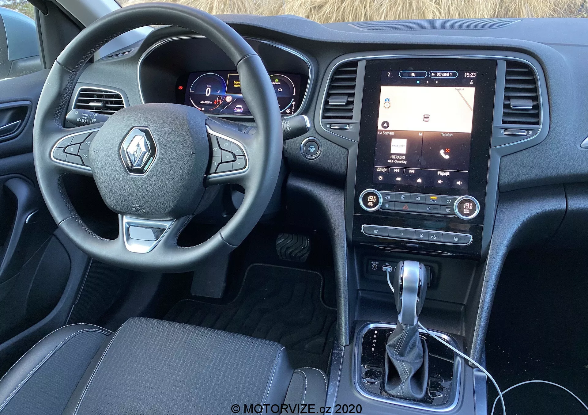 Interiör av en Renault Megane 2022 från förarens sätesperspektiv, vilket visar ratten med Renault-logotyp, digital instrumentpanel och en vertikalt orienterad pekskärms-infotainmentsystem på mittkonsolen. Detaljerad vy inkluderar knappar för olika funktioner på ratten, upplysta fordonsmätare, automatisk växelspak och minimala fysiska kontroller under infotainmentskärmen.