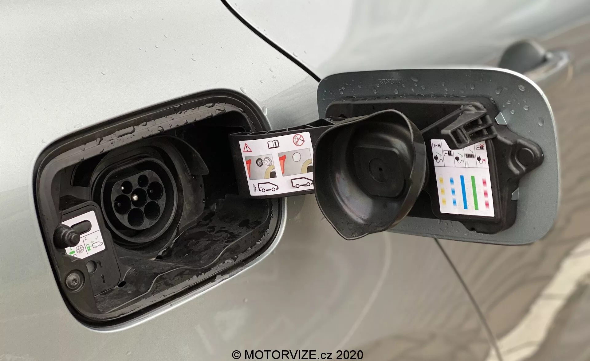 Närbild av laddningsområdet på en elbil (EV) från en diagonal vinkel, som fokuserar på den öppna laddporten som avslöjar kontakternas konfigurationer. Bilens kaross runt bränslekapsområdet är synlig, vilket tyder på att det är ett ljusfärgat fordon. Två separata kontakttyper visas, vilket tyder på stöd både för AC- och DC-laddning, där en liknar en typ 2-kontakt för AC och den andra en CCS-kontakt för DC-snabb laddning. Märkningen runt portarna inkluderar symboler för laddningstyper, spänningsinformation och en klistermärke med en QR-kod och färgade staplar. Laddportens dörr är fjäderlastad med synlig smuts och vatten, vilket indikerar regelbunden användning och exponering för väder. Bilden är tagen något nedåt mot kontakterna.
