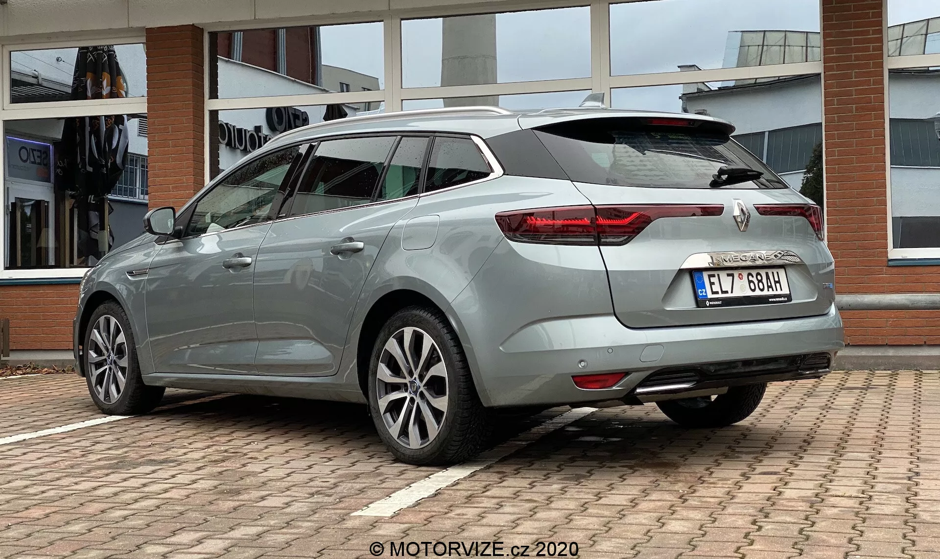 Trebaksvy bakifrån av en Renault Megane E-Tech 2022, som lyfter fram dess förlängda taklinje och bakluckedesign. Bilden visar distinkta LED-bakljus, silverfärgade legeringshjul med flerkekig design, sidokjolar, bakruteviskar, högt monterat bromsljus integrerat i bakspoilern och en hajfensantenn. Bilen har en ljus metallicblå/grå glansig finish med märkes- och modellmärken på baksidan. Positionerad på en parkeringsplats med en byggnadsfasad i bakgrunden, sedd från en låg vinkel för att betona bilens designelement.