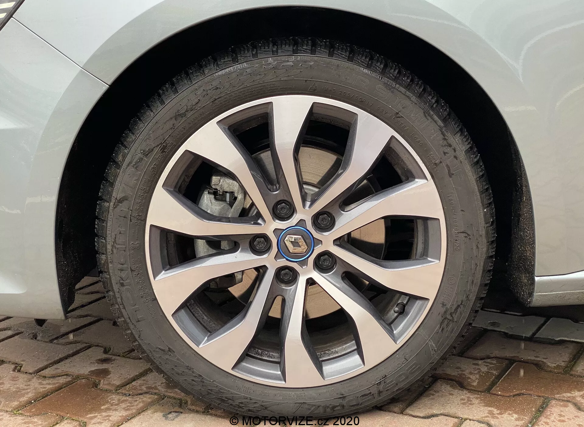 Närbild av framhjulet och däcket på en 2022 Renault Megane E-Tech från en sid- och något framåtvinklad perspektiv. Bilden framhäver den sofistikerade flerekrade designen med silver- och mörkgråa finishar, detaljerade slitbanemönster på däcket, en mittkåpa med varumärkets logotyp, synlig bromsskiva och bromsok och en liten del av den lackerade skärmen. Bilen står parkerad på en mark med fiskbensmönstrad beläggning i dagsljus.