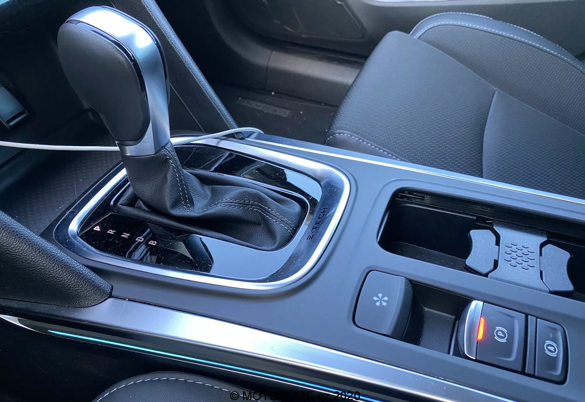 Renault Megane E-Tech 2022 mittkonsolinteriör med fokus på växlingsområdet, med en automatisk växelspak täckt av läder, körlägeskontroller, elektrisk parkeringsbromsknapp, mugghållare, styrplatta eller joystick för multimedia/navigeringssystem, litet förvaringsutrymme troligen med USB-portar och perforerad läderklädd sitskant. Högblank svart plast och metalldetaljer, vilket indikerar en modern och teknologiorienterad bil.