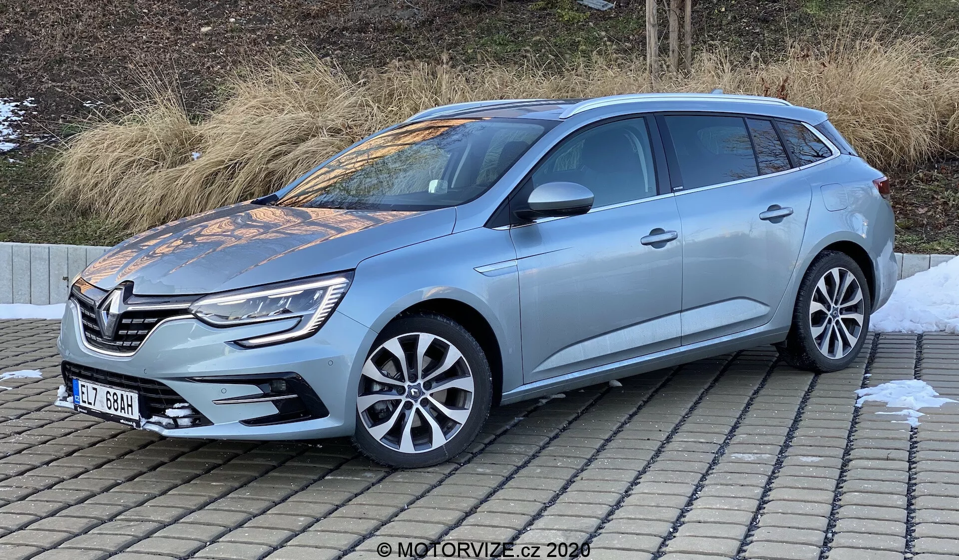 Bilden visar en ljusblå Renault Megane-e-Tech 2022 kombi avbildad från en trekvarts-vy framifrån, vilket ger en omfattande vy av både framsidan och sidan av fordonet. Den distinkta grillen och emblemet flankeras av eleganta strålkastarenheter, förmodligen med LED- eller xenonbelysning, kompletterat med integrerade dimljus och parkeringssensorer på främre stötfångaren. På sidan syns lättmetallfälgar och takräcken, vilket antyder nytto- och lastkapaciteter. Sidobackspeglarna kan ha integrerade blinkers eller dödvinkelövervakning. Fotograferad i dagsljus på nära håll, framhäver bilden bilens texturer, ytbehandlingar och övergripande design med tydlighet.
