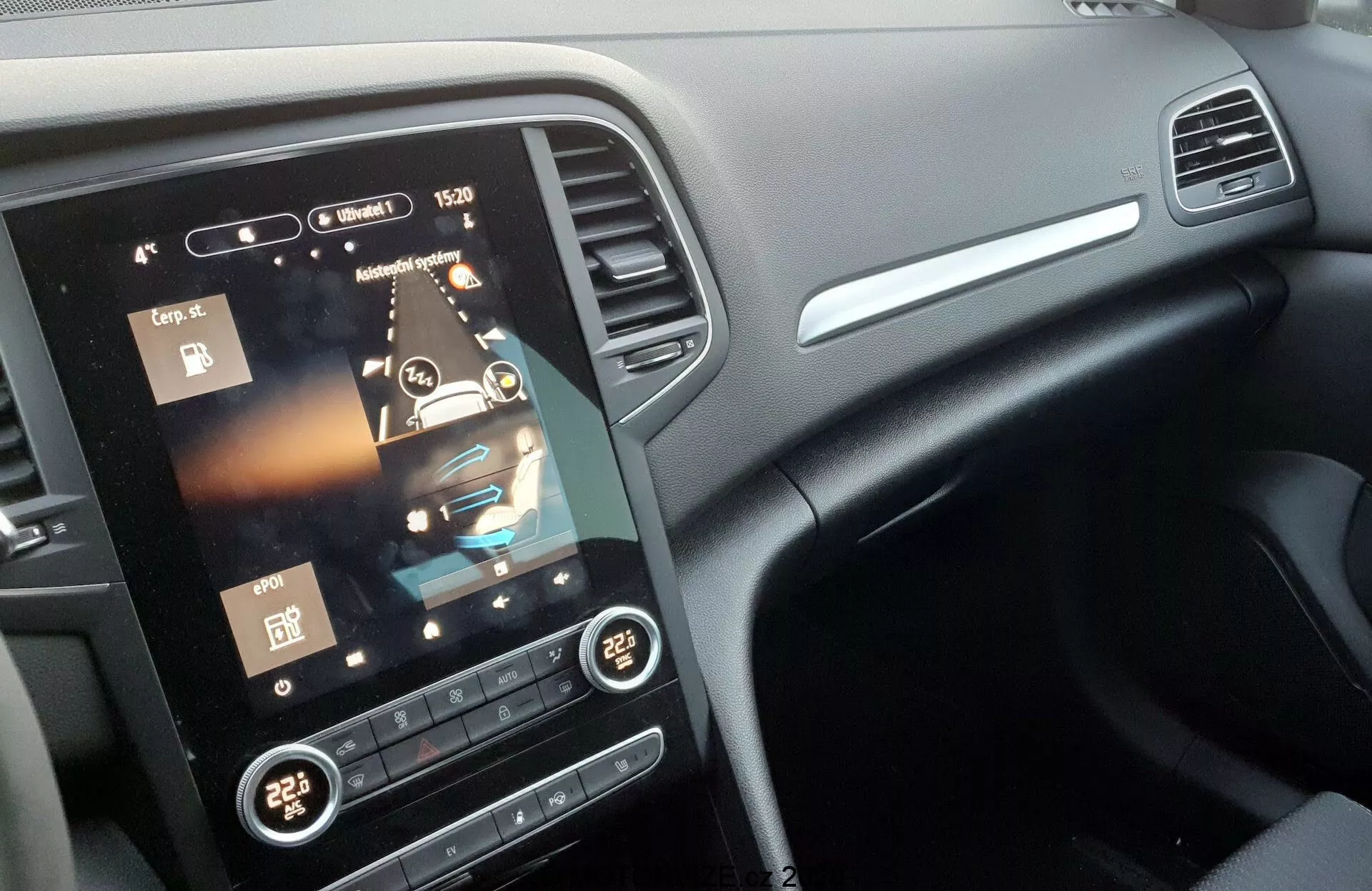 Närbild av centralkonsolen i interiören av Renault Megane 2022 (Megane-e-Tech) som visar den stora vertikala pekskärmen med infotainmentsystemet med ikoner för medieuppspelning, fordonsinställningar, klimatanläggning och navigering. Fysiska knappar och vred för HVAC-systemet under skärmen, dubbelzons klimatkontroll inställd på 22 grader Celsius. Mörk instrumentpanel med kontrasterande borstad metallfinish och ventilationsöppningar ovanför skärmen. Delvis syn av ratten från en sned vinkel, taget från framsätet på passagerarsidan.