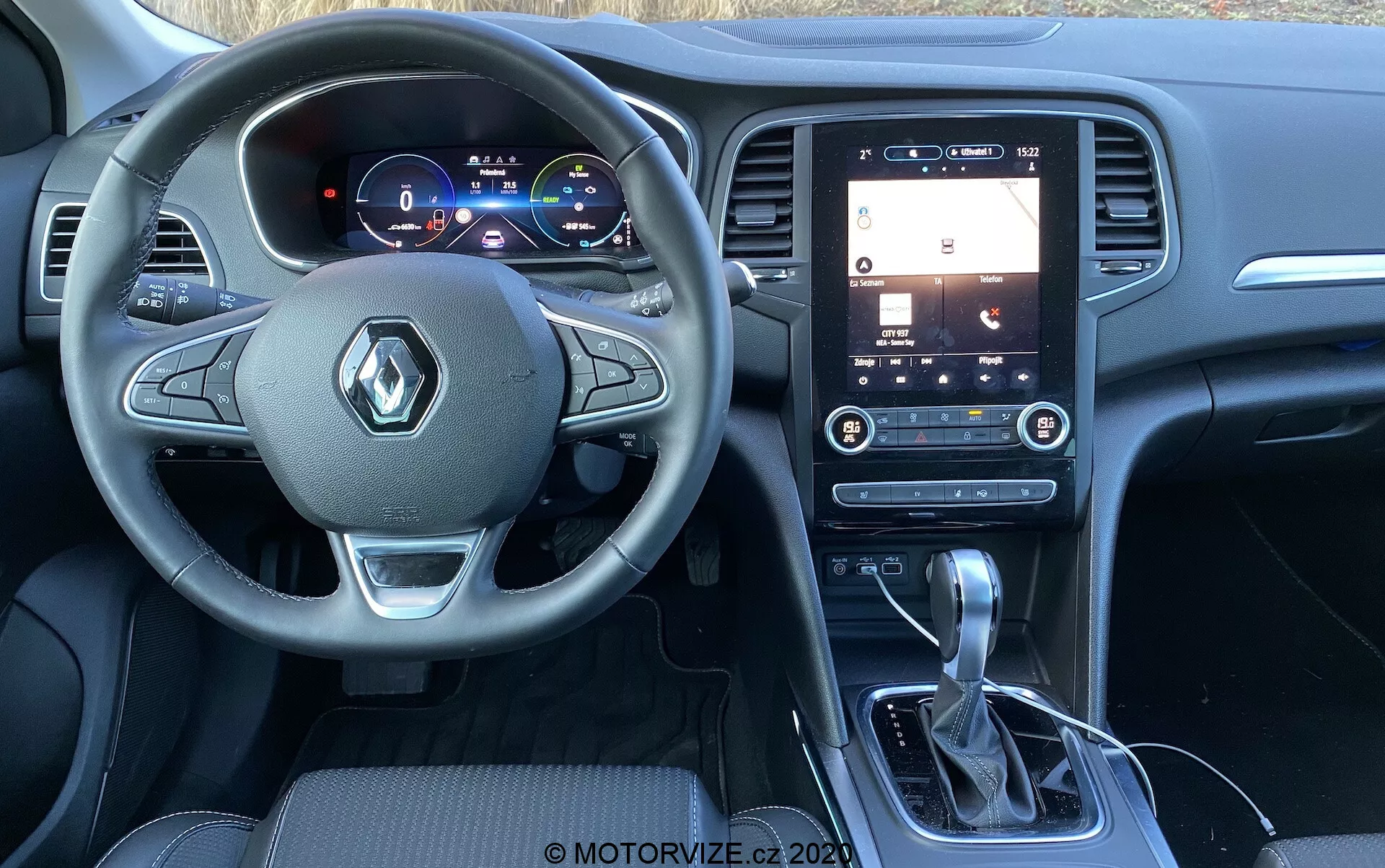 Interiörvy av 2022 Renault Megane-E-Tech fångad från passagerarens perspektiv, med fokus på ratten med Renault-logotyp och monterade kontroller, digitalt instrumentkluster som visar fordonsinformation, vertikalt orienterad pekskärm på mittkonsolen med infotainmentfunktioner som GPS-navigering och ljudinställningar, automatisk växelspak med läderskydd, mugghållare, vertikala luftventiler med klimatkontrollinställningar och premiummaterial i interiören inklusive mörka plaster och borstad metall runt växelspaken och ratten, med synliga kanter på läder- eller skinnliknande säten.