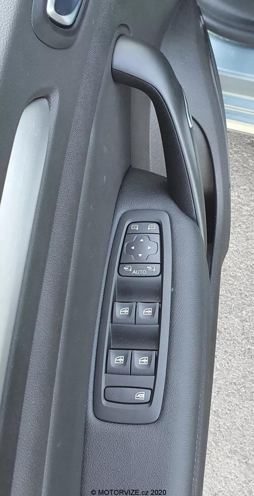 Närbild av kontrollpanelen på förardörrens insida i en Renault Megane E-Tech 2022. Bilden visar en vy från ovan av olika kontroller, inklusive strömfönsteromkopplare, fönsterlås omkopplare, vridbar omkopplare för inställning av sidospeglar, knapp för infällning av sidospeglar och eventuella minnesinställningar. Kontrollpanelen, tillverkad av robust mörk plast, framhäver fordonets moderna elektroniska kontrollsystem och komfortfunktioner.