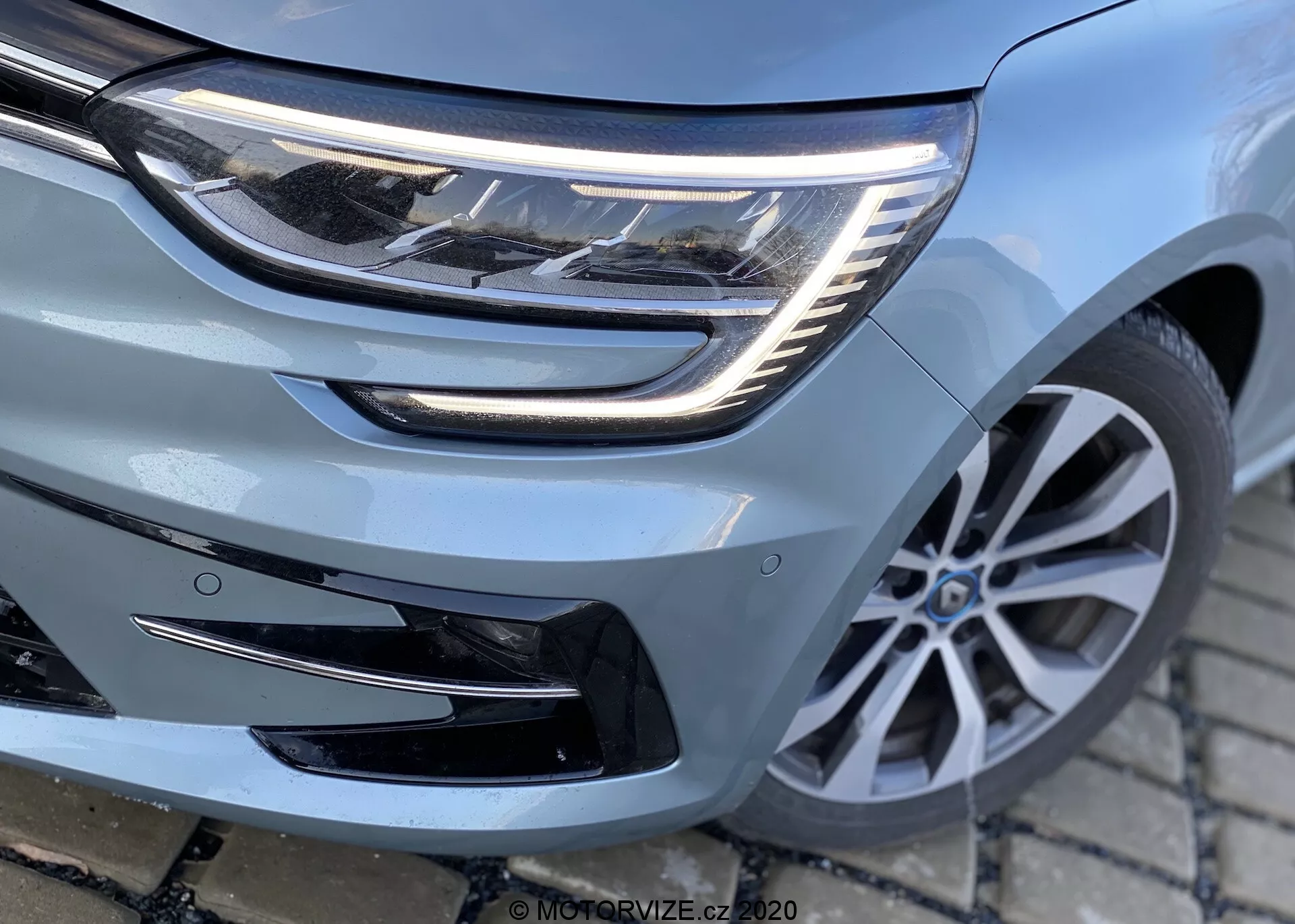 Närbild av fronten på Renault Megane 2022 Megane-e-Tech med fokus på strålkastare, grill och främre stötfångare, som visar LED-ljuselement, horisontell grilldesign och eventuell sensor för förarassistans. Med flerspoked lättmetallfälg och modern fordonsdesign från låg vinkel, något snett till vänster.