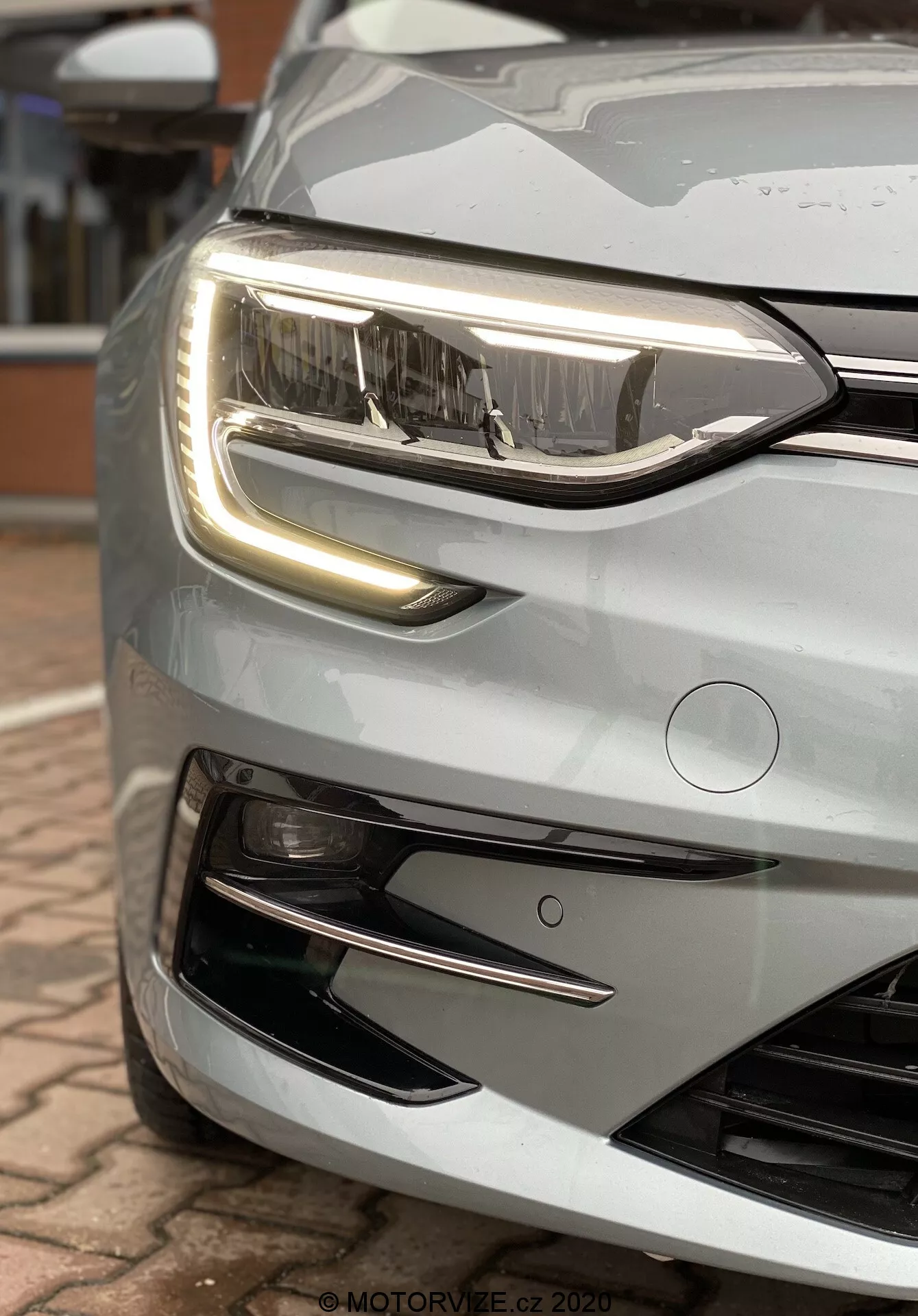 Närbild av fronten på Renault Megane E-Tech 2022, silverfärgad, fokuserande på modern LED-strålkastare och distinkt LED-dagsljus med synliga interna element. Bilden fångar den övre främre delen och framhäver grillen, motorhuvens hörn, den glänsande reflekterande ytan och dimljusens fördjupning. Fina detaljer inkluderar reflektioner, regndroppar på den rena ytan och en elegant vinkeldesign som betonar fordonets moderna estetik och avancerade belysnings- och säkerhetsfunktioner.