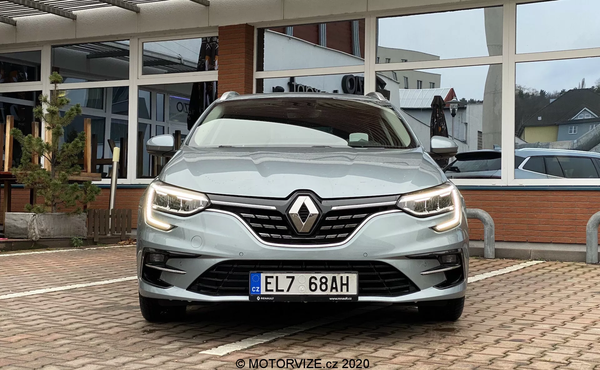 Närbild i vinkel framifrån av en silverfärgad Renault Megane E-Tech 2022, tagen från en låg vinkel nära grillen. Bilden visar bilens stiliga moderna LED-strålkastare, det framträdande Renault-emblemet på en grill med blandat mönster och motorhuvens aerodynamiska konturer. Den främre stötfångaren integrerar dimljus och luftintag, med betoning på både estetik och funktionalitet. Naturligt dagsljus med molnigt väder lyser upp fordonet, med ett asfalterat område och en glasfasad från en bilförsäljare i bakgrunden.