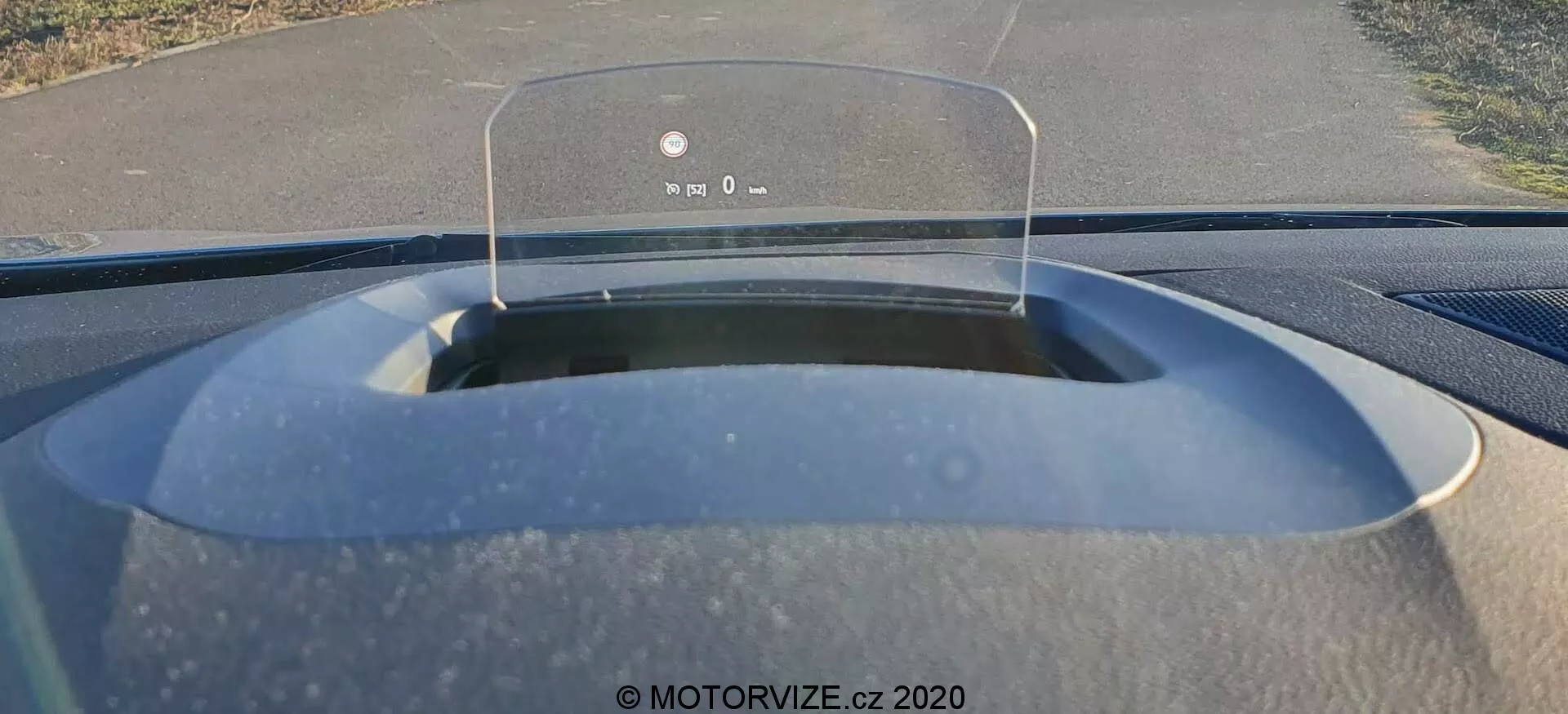 Invändig vy från förarsätet av en Renault Megane E-Tech 2022, med fokus på head-up displayen som visar viktig körinformation som hastighet och navigation på en transparent skärm ovanför instrumentbrädan. Vindrutan visar en enkelspårig väg med vegetation på båda sidor, vilket indikerar en förorts- eller lantlig miljö. Motorhuvens matta yta och synliga dammpartiklar antyder att bilen nyligen har använts eller behöver rengöras.