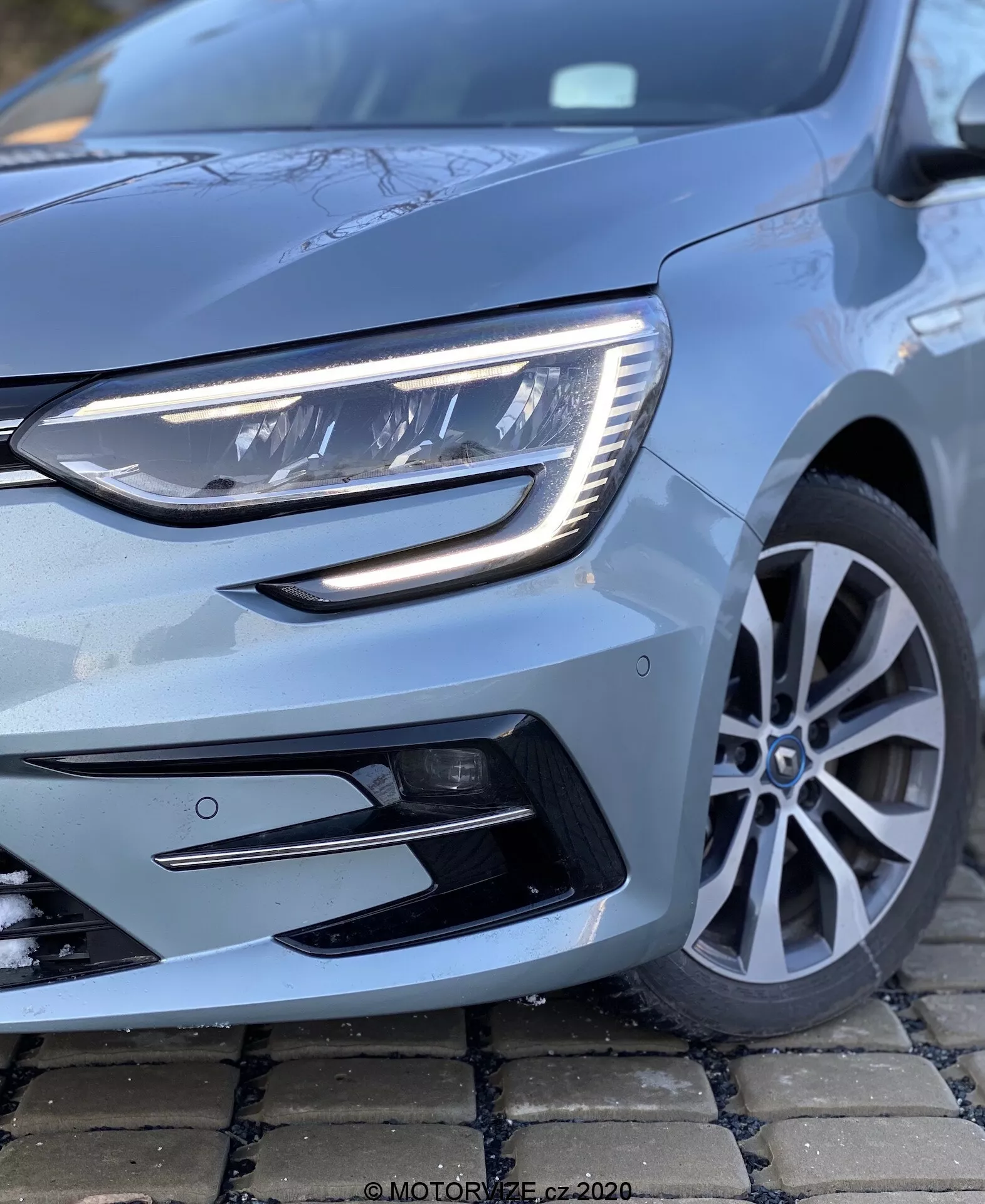 Närbild av Renault Megane 2022 (Megane-e-Tech) från låg vinkel med fokus på vänstra främre kvadranten. Bilden visar en samtida strålkastare med LED-element och en elegant dagsljuslampa. En del av den främre grillen och stötfångaren med svart accentuerade luftintag är synlig, tillsammans med den snygga fler-ekrade aluminiumfälgen och en metallic ljusblå lackfinish som speglar fordonets skick.