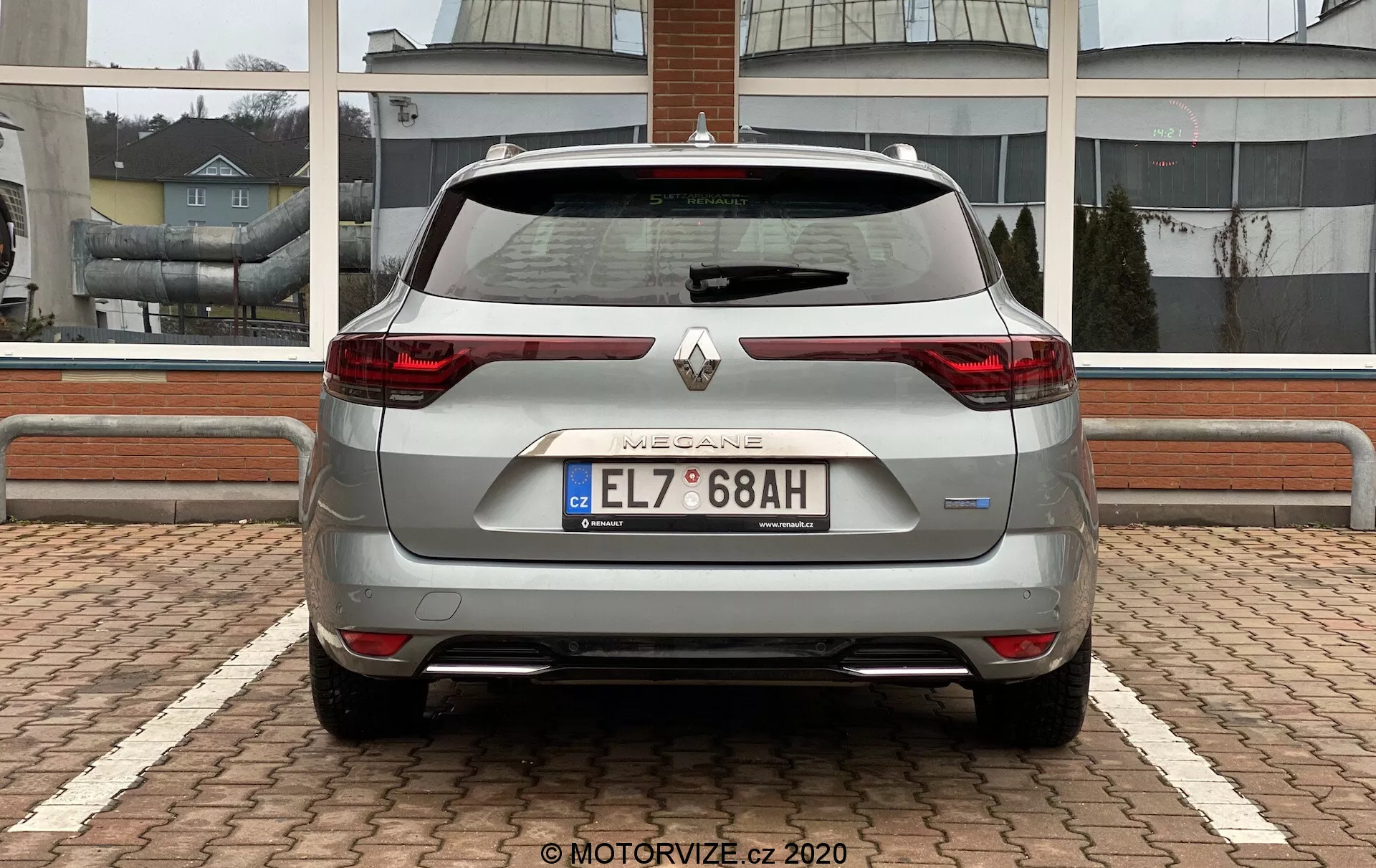 Bakifrån vy av en silverfärgad Renault Megane E-Tech 2022, med C-formade LED-bakljus, centralt Renault-emblem under inskriptionen 'MEGANE', europeisk registreringsskylt, parkeringssensorer, yttre reflexer, bakrutesviskare, mjukt sluttande taklinje som slutar med takmonterad spoiler, hajfenneantenn, ren bakestetik med dold avgas och delvis synliga aluminiumfälgar. Bilen är parkerad på ett fisksbensmönstrat markbeläggning med en kommersiell eller bostadsbyggnad i bakgrunden.