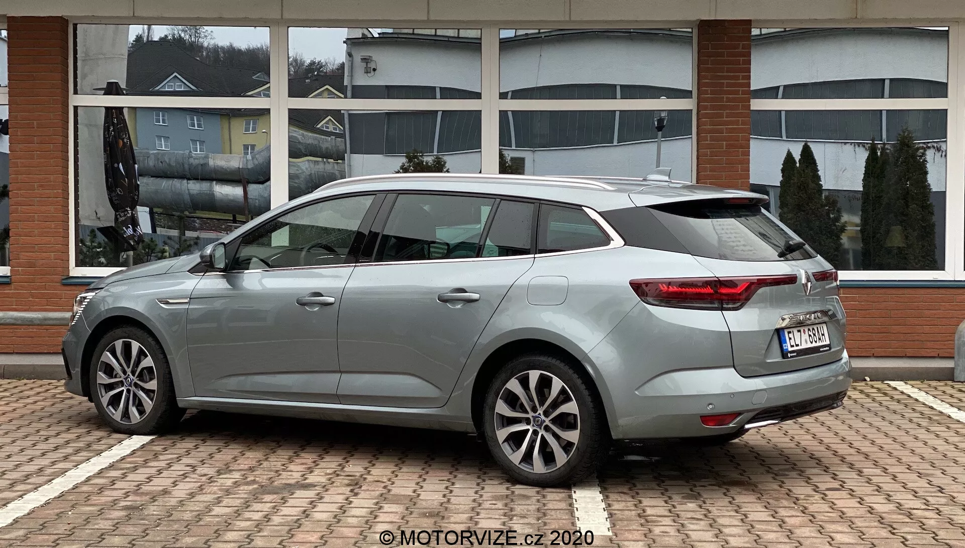 Sidoprofil av en silvergrå Renault Megane 2022 kombi, parkerad på en yta med tegelmönster, med slanka konturlinjer, sidospeglar och dörrhandtag i karossens färg, flerekerfälgar i lättmetall, distinktiva bakljus som sveper runt hörnen, bakrutespolare, högmonterat bromsljus och takspoiler. Bilen är placerad mot en byggnad med stora fönster som speglar den molniga himlen.
