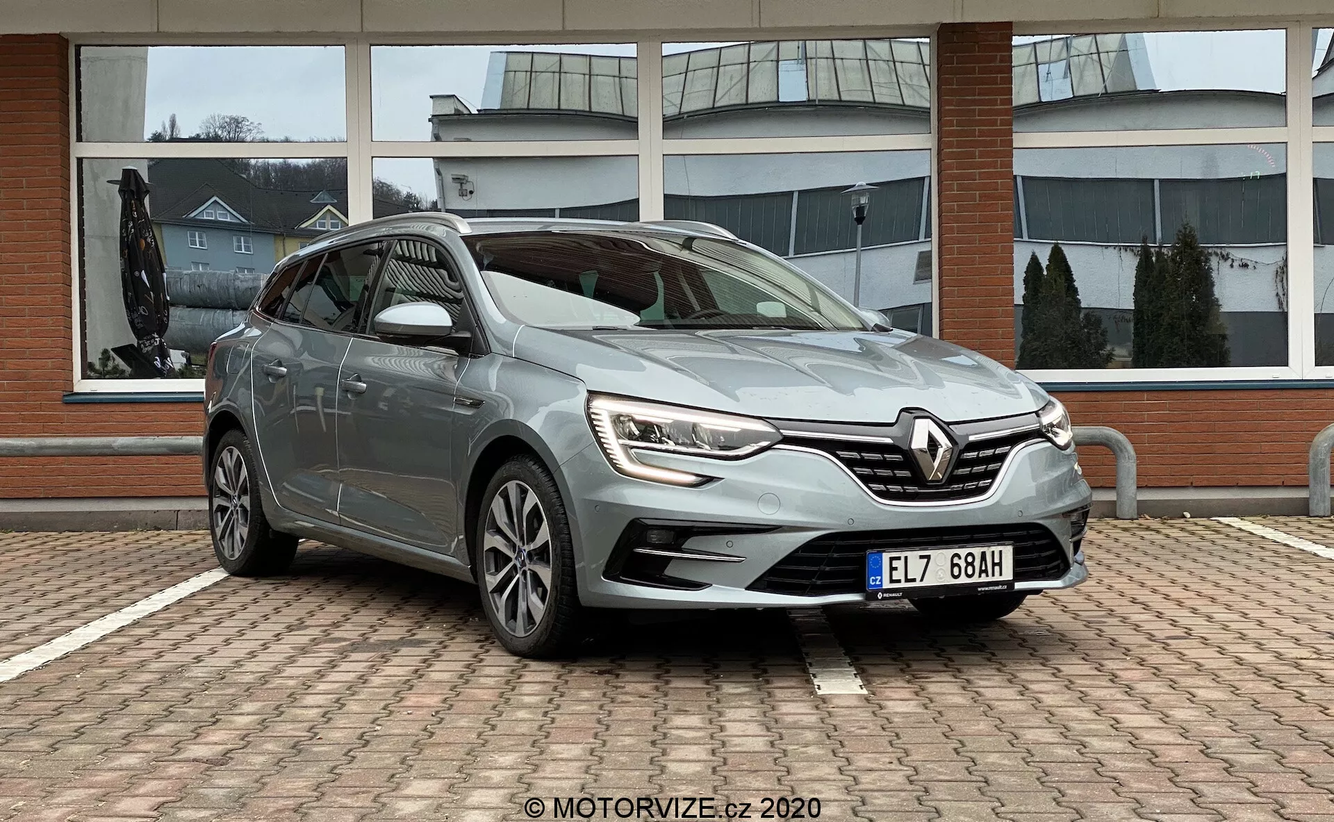 Främre tre-kvarts vy av en Renault Megane 2022 (Megane E-Tech) halvkombi, parkerad på ett tegelbelagt område med en kommersiell byggnad med stora glasfönster i bakgrunden. Fotograferad från en framvinklad förarposition, lyfter bilden fram bilens LED-dagljus, intrikata grilldesign med Renault-logotypen, skulpterade sidolinjer, flerspakiga lättmetallfälgar och sidospeglar med integrerade blinkers. Mulet väder ger mjukt, diffust ljus som förbättrar synligheten för designfunktionerna.