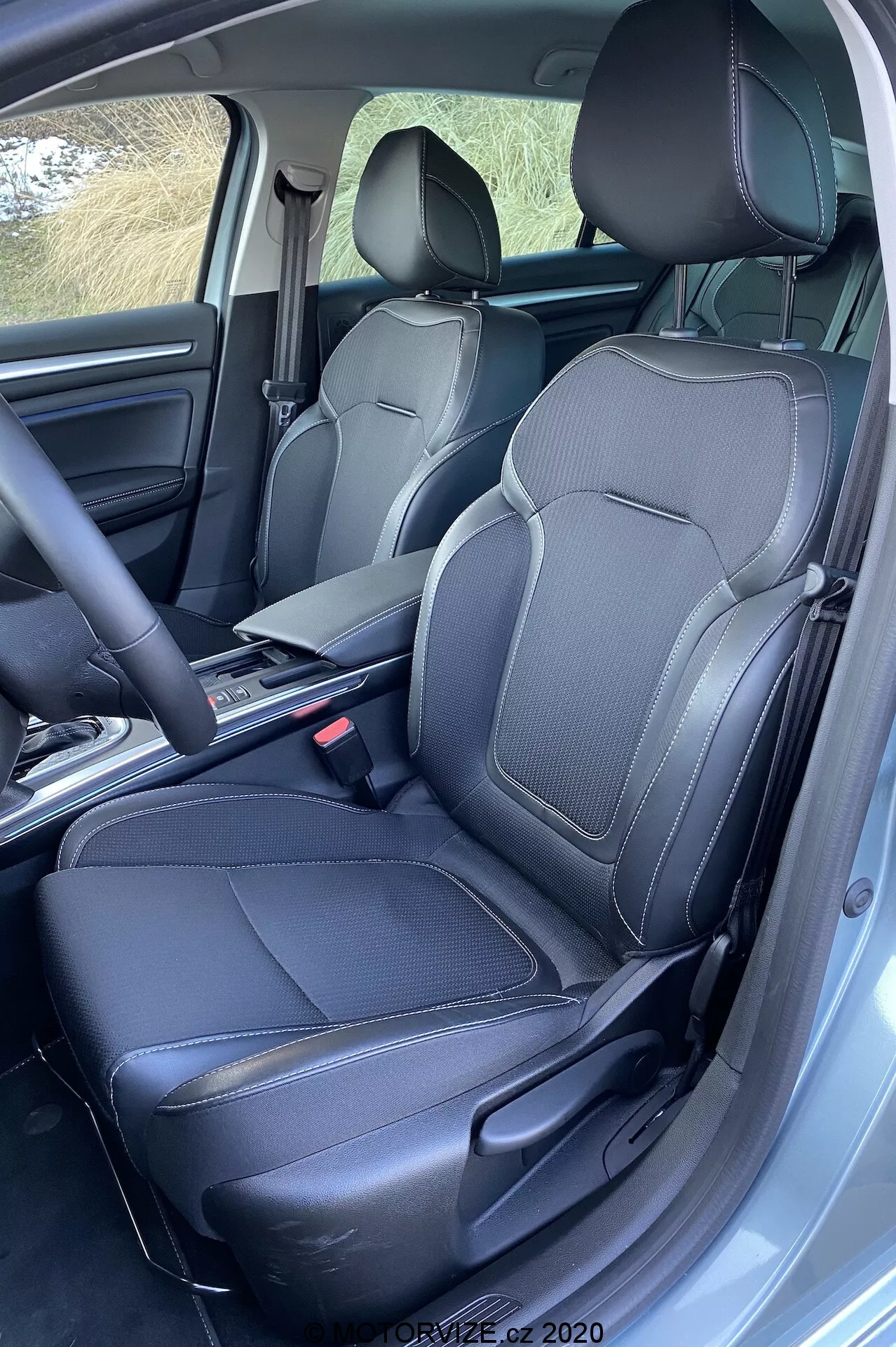 Interiör av Renault Megane 2022, främre passagerarområdet med svarta lädersäten med detaljmönstrad söm, nackstöd och sidostöd. Central konsol med otydlig växelspak och nedfälld armstöd, vilket ger förvaringsutrymme. Synliga säkerhetsbälten och förankringsmekanism, heltäckningsmattor med specialanpassade mattor. Förarsidan dörr öppen, något förhöjd vinkelvy som visar en premiumfinish.