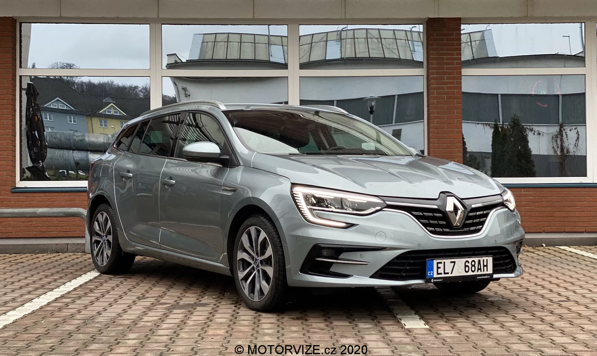 Trekvarts framifrån vy av en stillastående Renault Megane E-Tech 2022, som visar den eleganta främre grillen med LED-dagljus, dimljus och aerodynamisk stötfångare. Sidprofilen framhäver förardörrs paneler, fönster, speglar med blinkers och flerekrade lättmetallfälgar. Metallfärg, blank finish, parkerad på ett mönstrat tegelyta, förmodligen på återförsäljarens förgård. Naturligt ljus förbättrar det rena och välskötta utseendet.