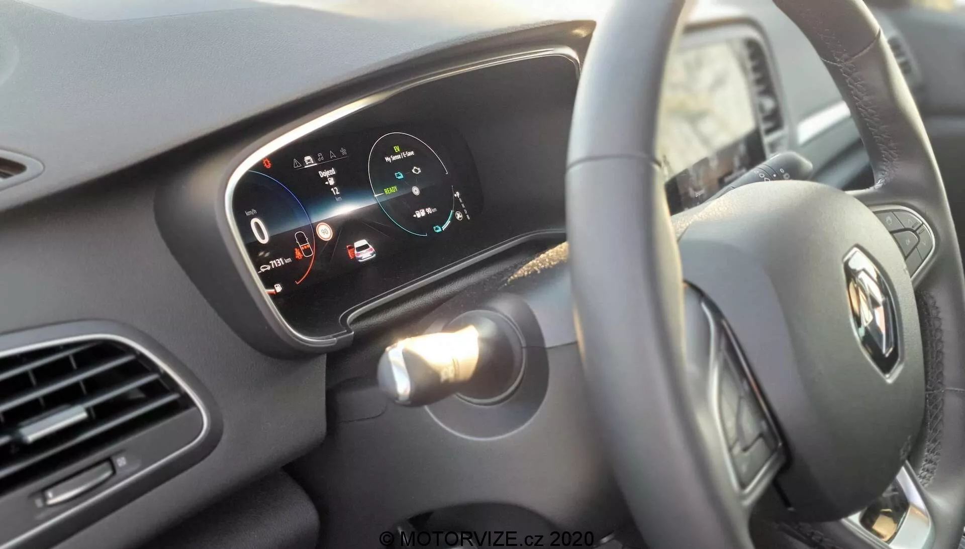 Interiörvy av Renault Megane E-Tech 2022 från förarens perspektiv, med fokus på den centrerade ratten med Renaults logo och den digitala instrumentpanelen bakom. Ratten har en enda spak synlig till vänster för blinkers och eventuell kontroll av strålkastare. Den digitala skärmen visar en hastighetsmätare som läser 0 km/h och en effektmätare med zoner markerade 'charge', 'eco' och 'power', vilket indikerar att fordonet är elektriskt eller hybrid. Varningslampor och ikoner för batterinivå, varning för öppen dörr och andra funktioner är synliga. Till vänster syns en del av infotainment- eller navigationssystemet, delvis täckt av ratten. Den minimalistiska interiördesignen med horisontella linjer över instrumentpanelen och ventilationsöppningar betonar bilens bredd. Fokusdjupet framhäver rattens emblem och den digitala skärmen, vilket betonar det avancerade digitala gränssnittet för instrumentpanelen.