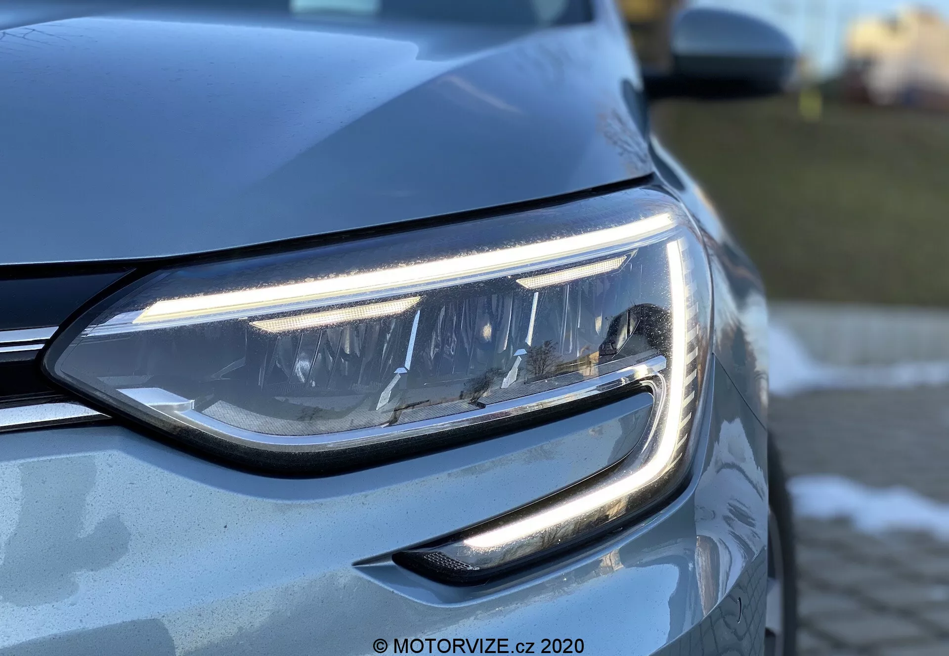 Närbild på förarens sidas strålkastare på en Renault Megane 2022 (Megane-E-Tech) tagen i en sned vinkel framifrån. Betonar de eleganta LED-dagsljusen och projektorlampan, med en glimt av det moderna gallret och den metalliska lacken som reflekterar ljuset.