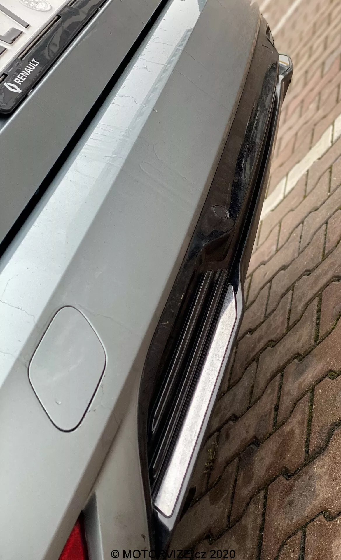 Närbild av den högra bakre delen av en Renault Megane E-Tech 2022, med fokus på bakljusen med LED-lampor, kanten av bagageluckan med integrerad spoiler och en liten del av bakre stötfångaren med möjligt parkeringssensor eller backkamera. Fordonets metalliska lack är ren och speglar omgivningen, placerat på en tegelstensbelagd yta.