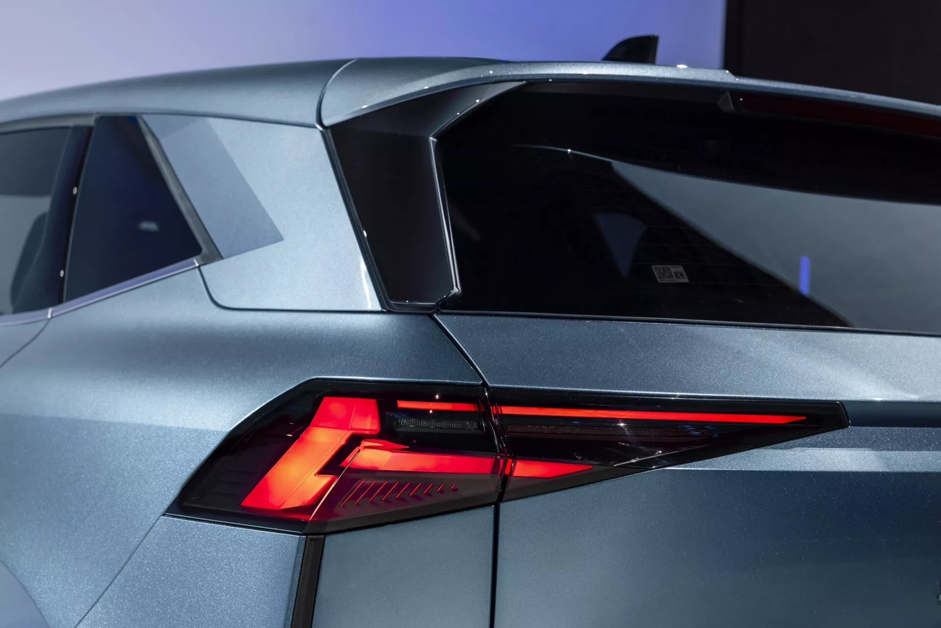 Närbild av en modern Renault Symbioz 2024 (1:a generationen) från en trekvart bakre vinkel. Bilden framhäver den skarpa, vinklade designen av LED-bakljusen, den sluttande bakrutan och den distinkta taklinjen med en liten spoiler. Karossen visar futuristiska geometriska former i en metallisk silver/grå finish. En badge eller emblem är synlig på vänster sida av bagageluckan, vilket antyder bilens modell eller märke. Andra detaljer inkluderar en sektion som liknar en bränslepåfyllningslucka på den vänstra bakre panelen.