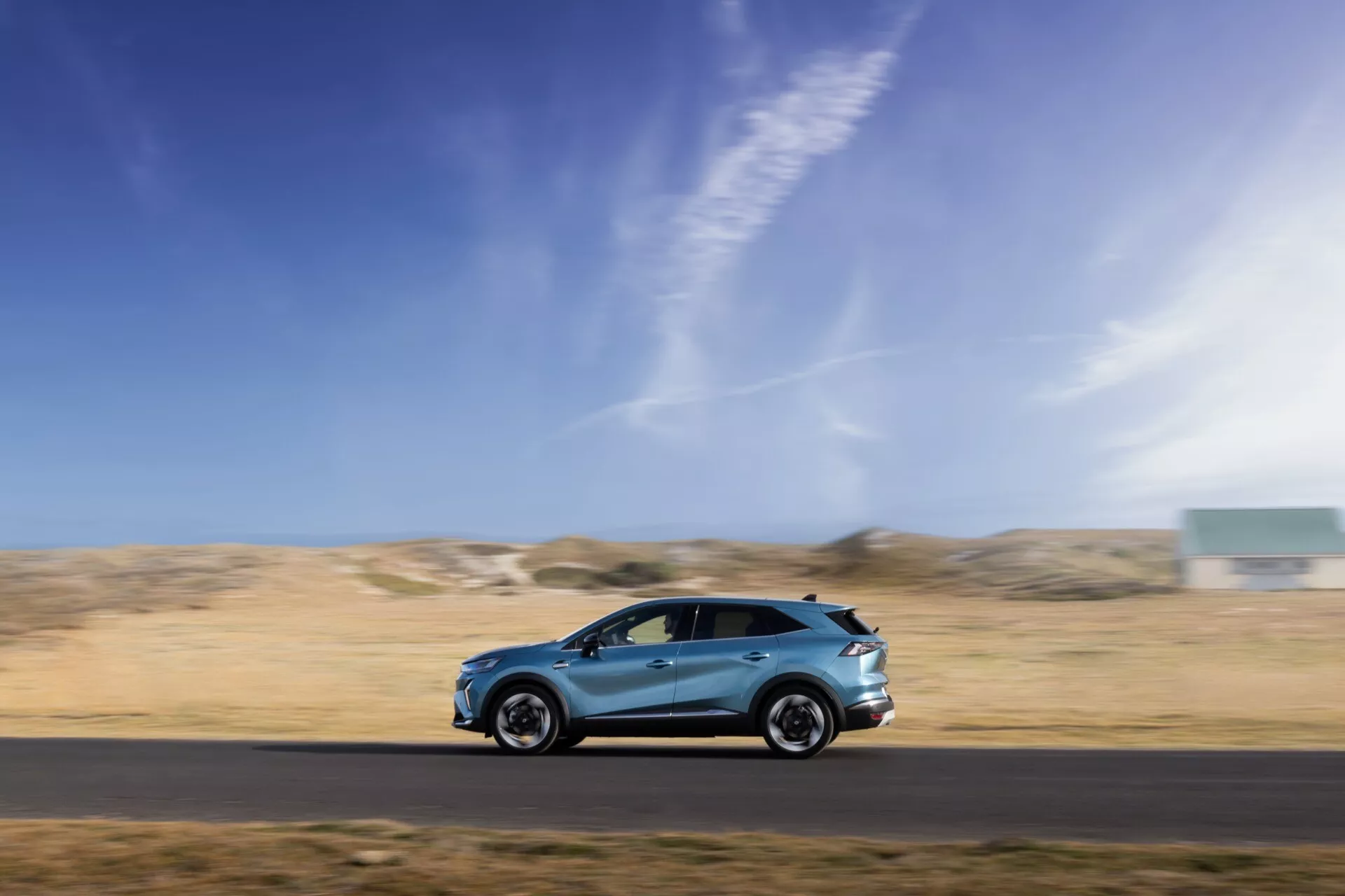 Dynamisk sidoprofilbild av den rörliga Renault Symbioz 2024 1st generation, fångad med panoreringssteknik mot en suddig bakgrund, som visar dess eleganta aerodynamiska design, legeringshjul, sidospeglar, integrerade dörrhandtag och högkvalitativ lackfinish. Fordonet är placerat i den nedre tredjedelen av ramen mot en blå himmel och ett kargt landskap, vilket betonar hastighet, rörelse och bilens förmåga i olika miljöer.
