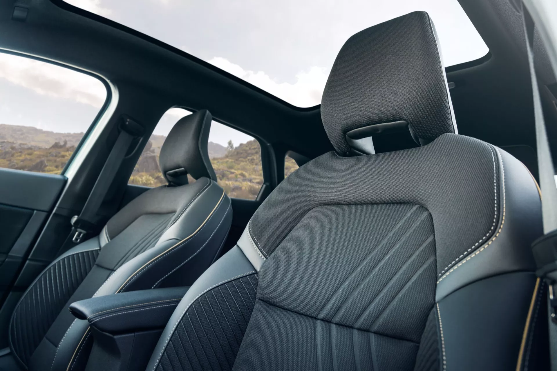 Interiör av Renault Symbioz 2024, med fokus på framsätena sett från baksätet. Ockuperade moderna designade säten med premium tyg- eller läderklädsel, detaljerade sömmar och ergonomisk formgivning. Integrerade nackstöd, sidoskålning och mörkfärgad klädsel antyder en kombination av komfort och sidostöd. Sidorutor och B-stolpar är synliga, som antyder säkerhetsfunktioner som sidoridåairbags. Naturligt ljus kommer in i kupén, vilket indikerar dagtid.