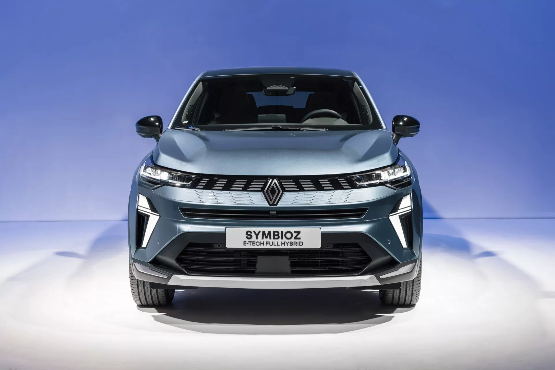 En närbildsfrontalbild av Renault Symbioz 2024 (1:a generationen) tagen från en något låg vinkel. Bilden betonar den eleganta grillen med en stiliserad 'R'-emblem, vinklade LED-strålkastare och en aerodynamisk motorhuv. Bilens metalliska ljusblå lack och blanka yta framhävs mot en ren, graderad blå bakgrund. Texten 'SYMBIOZ' på registreringsskyltens område antyder en modern, kanske futuristisk design med funktioner som LED-belysning, potentiella autonoma system och avancerad anslutning eller eldrivteknik.