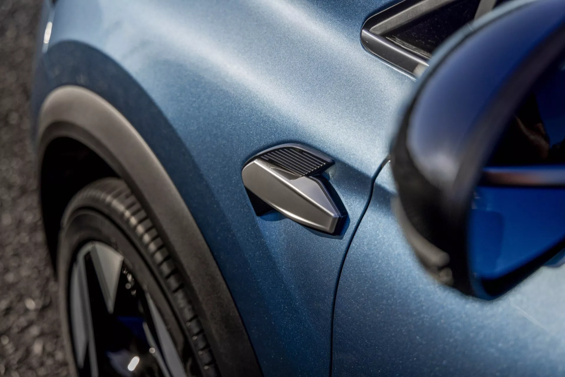 Närbild av en blå Renault Symbioz 2024 (1:a generationen), med fokus på sidopanelens kurvatur. Fotot framhäver en funktionell sidoventil, en del av ett flerspjälkat svart framhjul och den metalliska lackeringen på karossen. Den grunda vinkeln och selektiva fokuseringen framhäver bilens aerodynamiska och designmässiga detaljer, med en oskarp bakgrund.