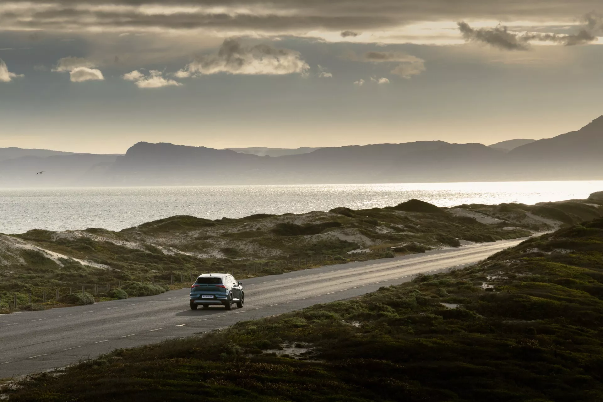 Högvinkelbild av en bil som kör på en slingrande kustväg med ojämn terräng och låg vegetation vid soluppgång eller solnedgång. Bilen, som verkar vara en SUV eller crossover, är placerad till vänster i ramen och rör sig åt höger. Scenen inkluderar ett lugnt hav, avlägsna berg och en liten segelbåt.