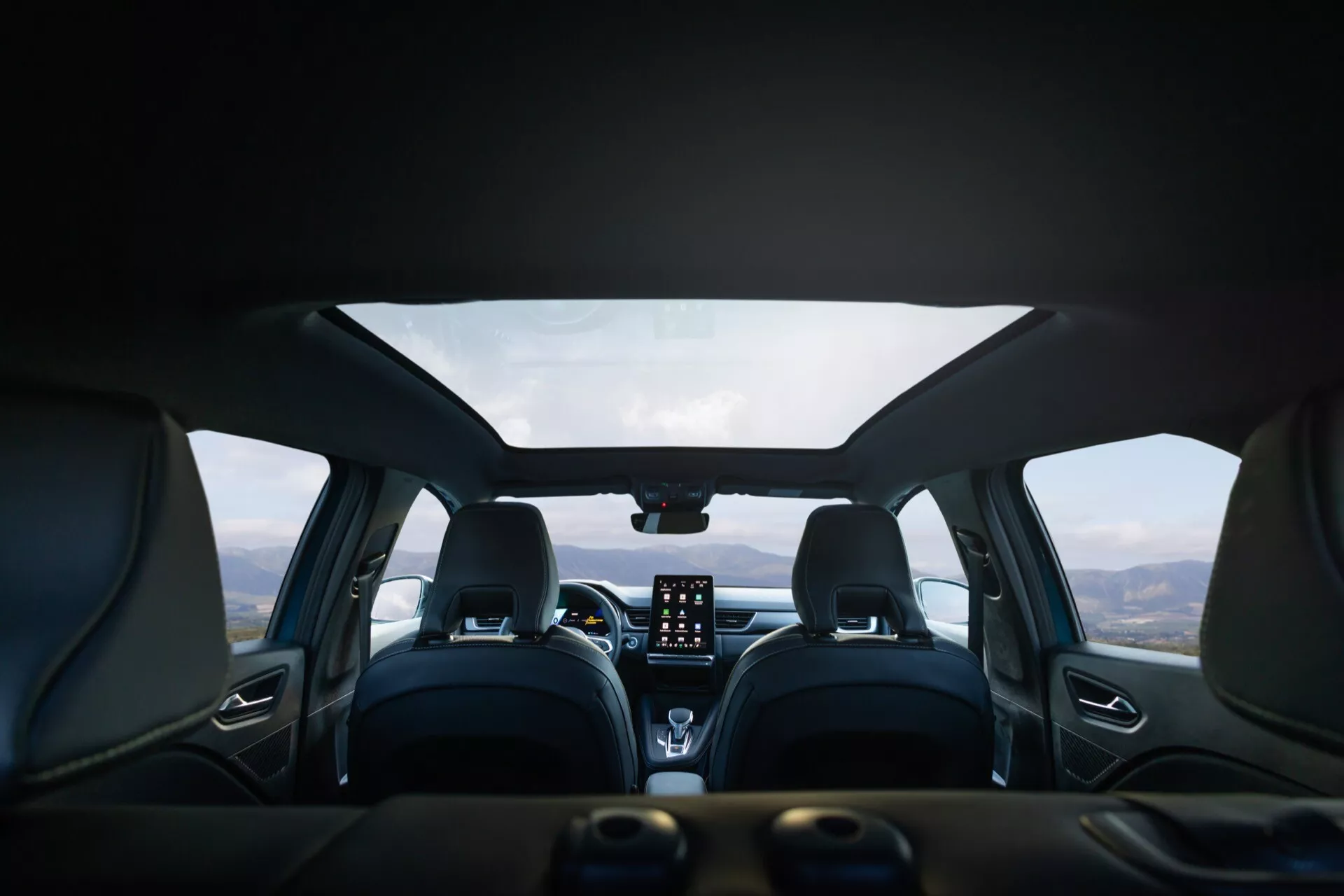 Interiörbild av Renault Symbioz 2024 (första generationen) från baksätets perspektiv, som visar en modern design med läderklädsel, distinkt sömnad och bekvämt formade framsäten med nackstöd. På mittkonsolen finns ett framträdande vertikalt pekskärmsinfotainmentsystem, klimatkontrollrattar och en digital display eller HUD ovanpå. Den multifunktionella ratten är delvis synlig. Genom vindrutan ser man ett bergslandskap under en mulen himmel, vilket antyder att fordonet står stilla på en pittoresk plats.